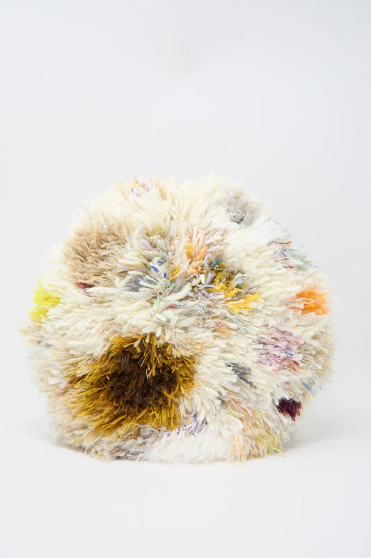 The Handmade Cushion in Multi XIII by Renilde De Peuter is a colorful, round pom-pom cushion featuring white, brown, yellow, and more on a light background. It's crafted using the Smyrna stitch technique with recycled yarns for an eco-friendly touch.