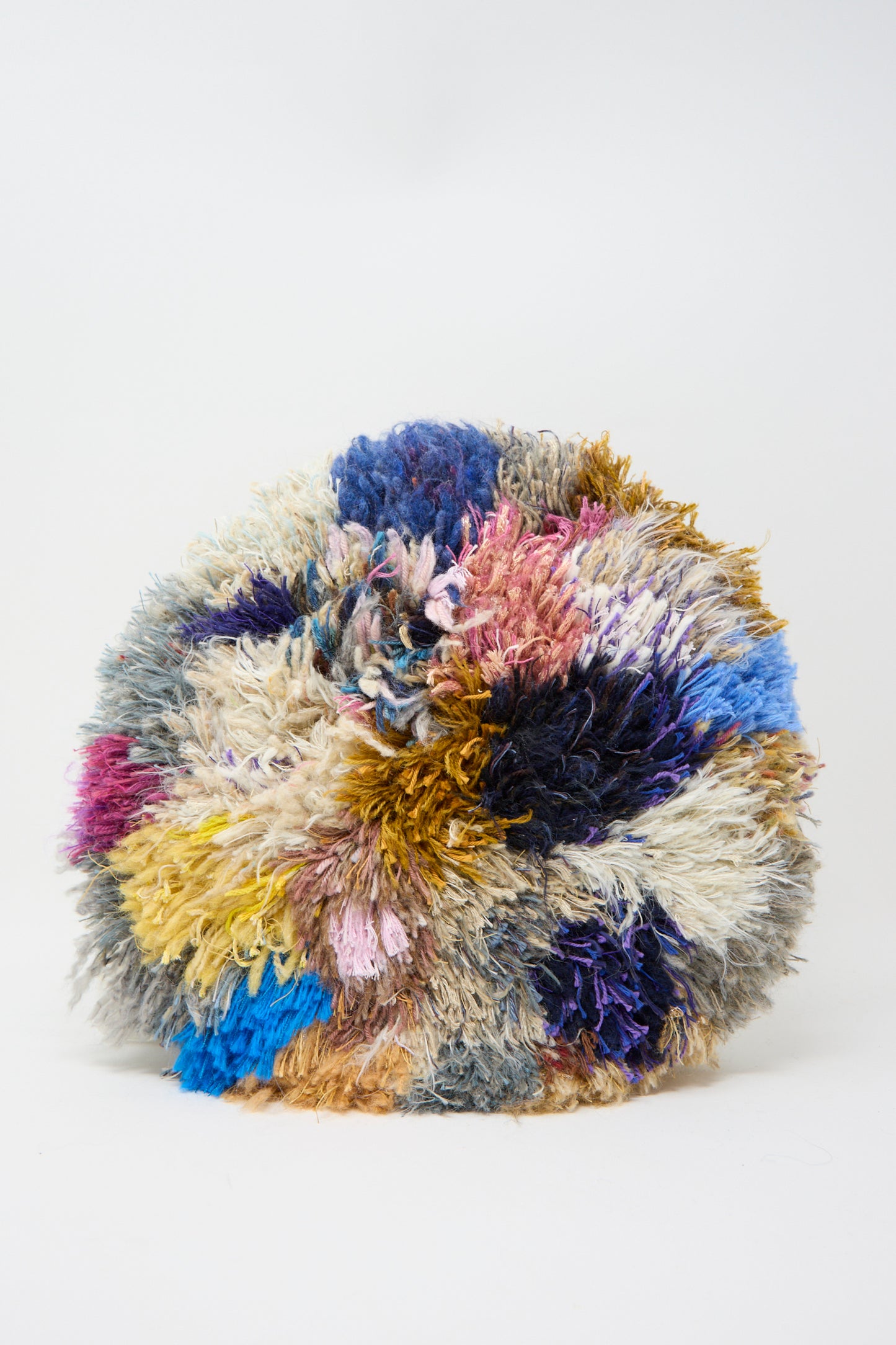 The Handmade Cushion in Multi XIV by Renilde De Peuter is a spherical, multicolored shaggy piece crafted with recycled yarns. It features blue, pink, yellow, and brown hues against a white background with intricate Smyrna stitching for an artisanal touch.