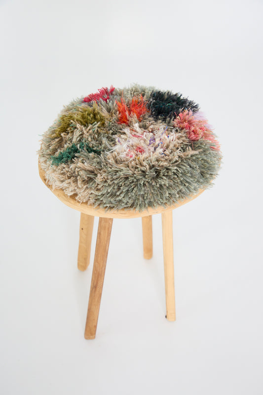 The Handmade Cushion in Multi XI by Renilde De Peuter sits on a wooden stool, offering a round, multicolored, shaggy cushion made from recycled yarns in green, red, and beige.