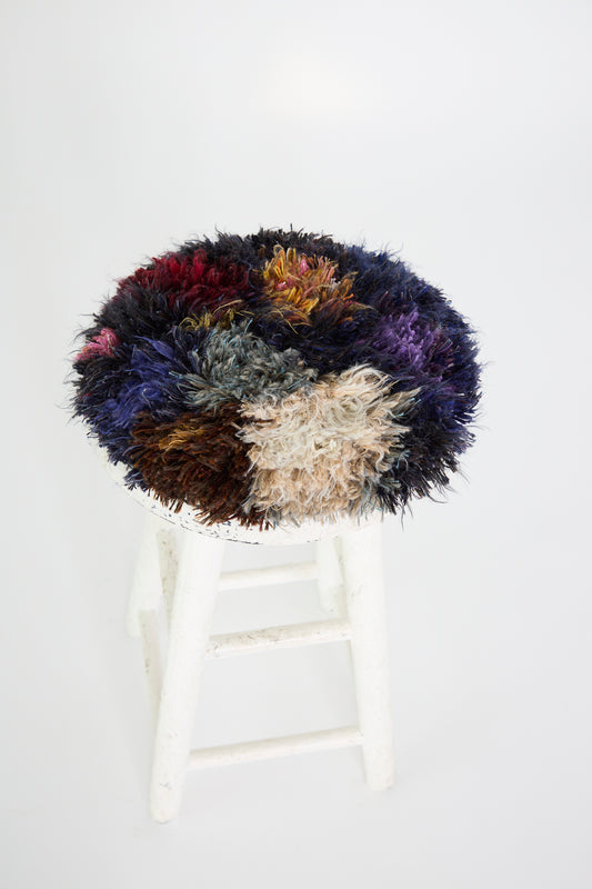 The white wooden stool showcases a "Handmade Cushion in Multi XII" by Renilde De Peuter, featuring a colorful, fluffy textured smyrna stitch made from recycled yarns against the plain white background.