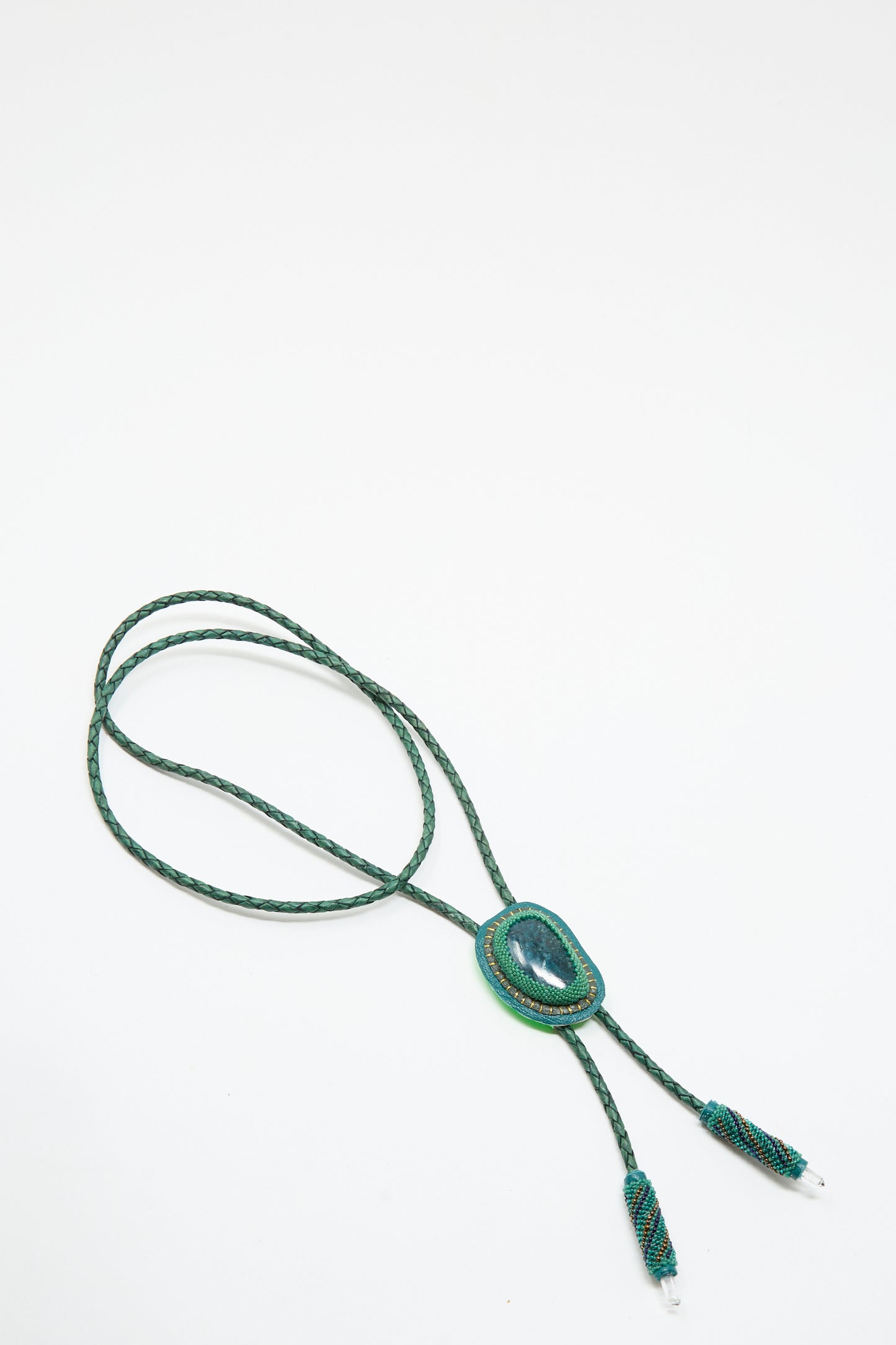 The Robin Mollicone Bolo Necklace in Apatite and Quartz features a green braided cord, a central oval quartz stone, and decorative metal tips, all shown against a plain white background.