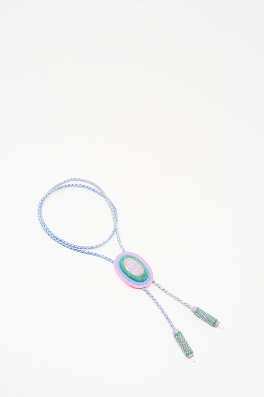 The Robin Mollicone Bolo Necklace in Kunzite and Quartz is a handmade blue braided piece from NYC, showcasing a pink and green oval centerpiece with decorative ends, shown on a white background.