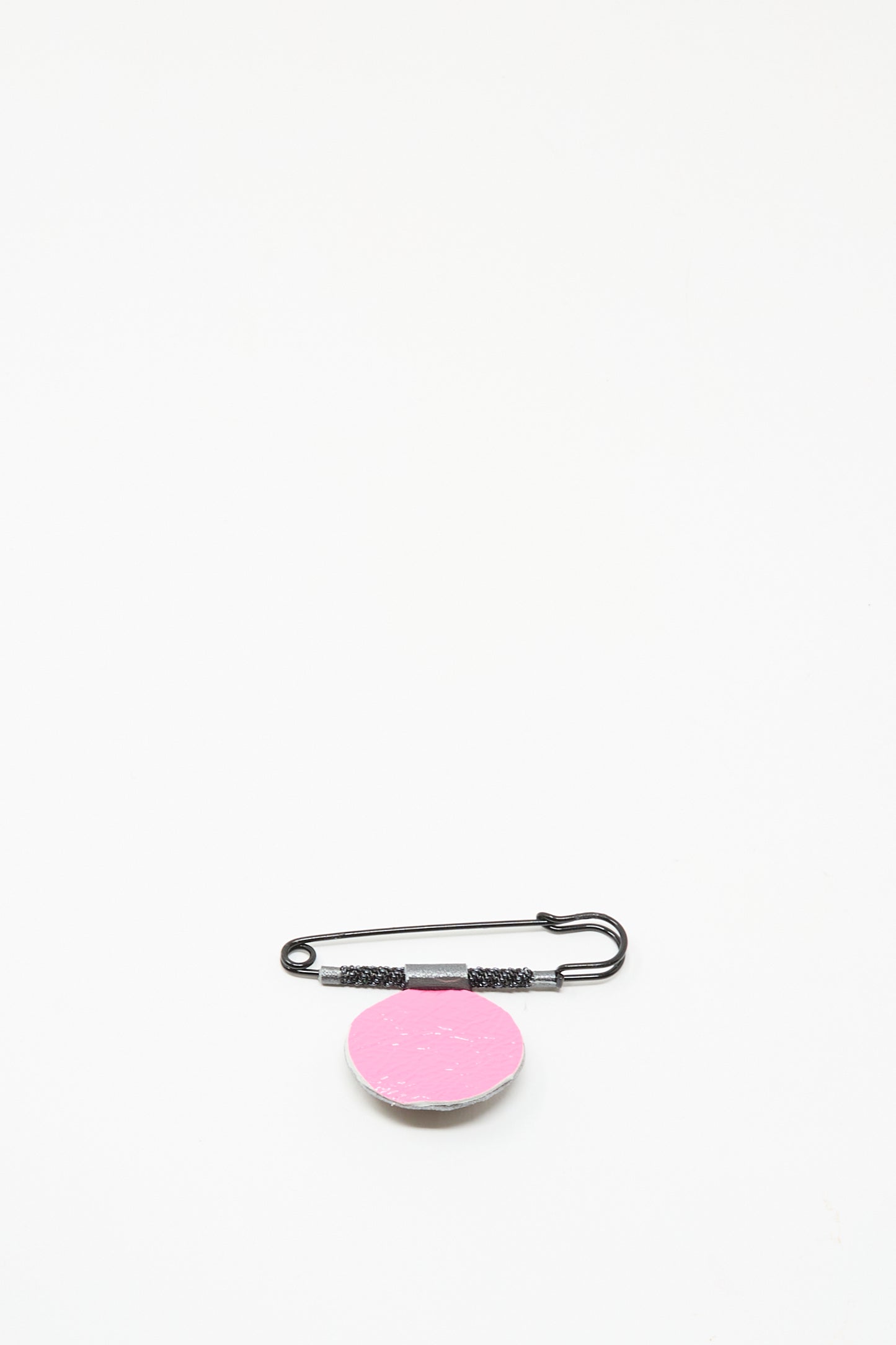 The Brooch in Onyx by Robin Mollicone features a black safety pin with a round, pink, textured disk. Handmade in NYC, it resembles a beaded brooch and is shown against a plain white background.