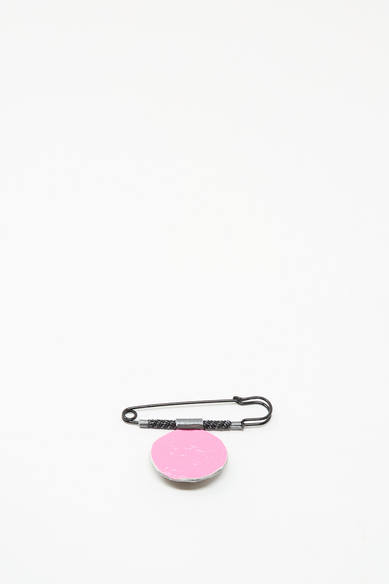 The Brooch in Onyx by Robin Mollicone features a black safety pin with a round, pink, textured disk. Handmade in NYC, it resembles a beaded brooch and is shown against a plain white background.