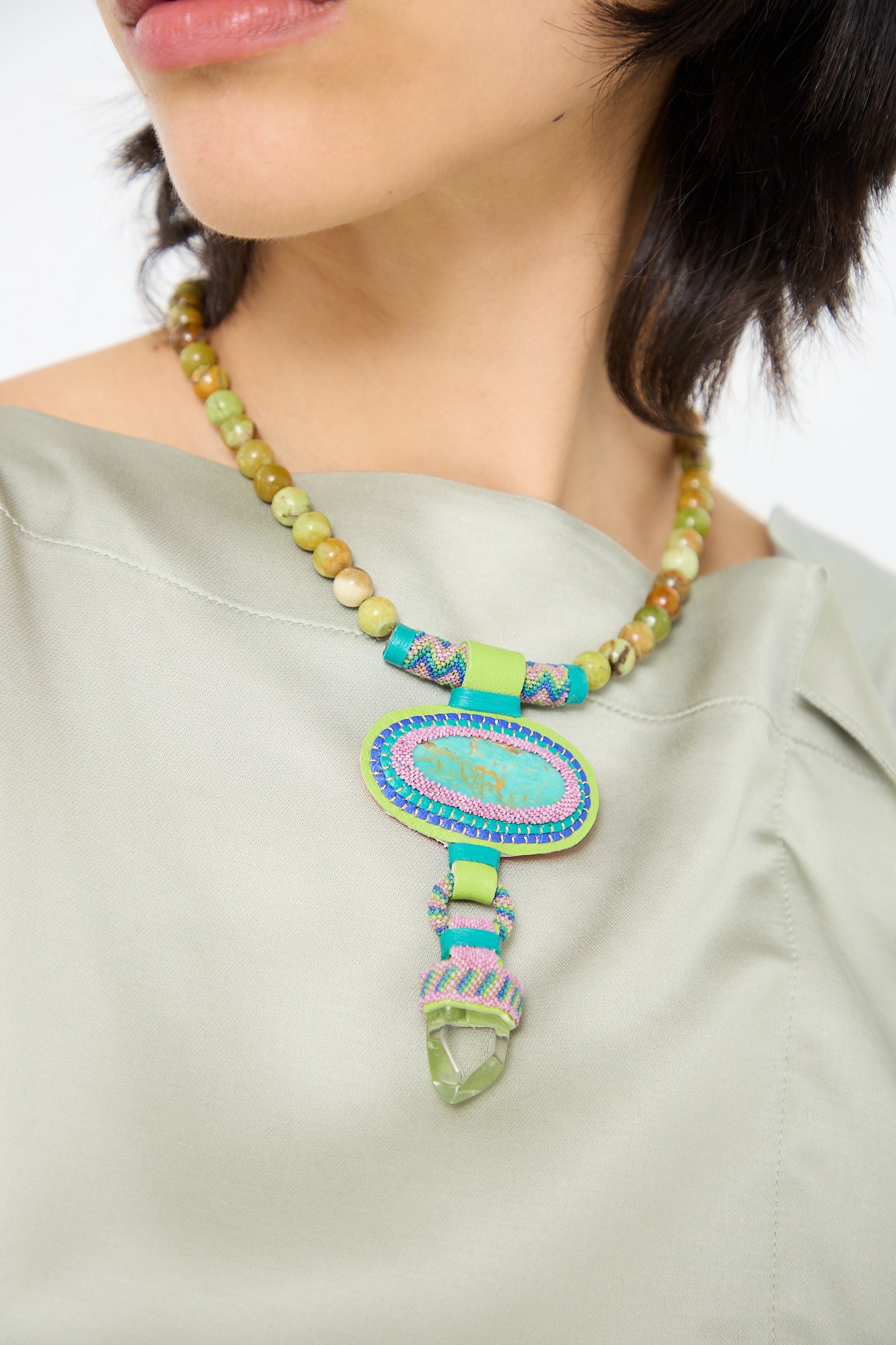 Close-up of a woman wearing a sage-green top and an intricate beaded necklace adorned with olive opal beads and a central turquoise stone, featuring green and yellow beads along the strand, the Double Stone Necklace with Crystal, Olive Opal Beads and Turquoise Stone by Robin Mollicone.