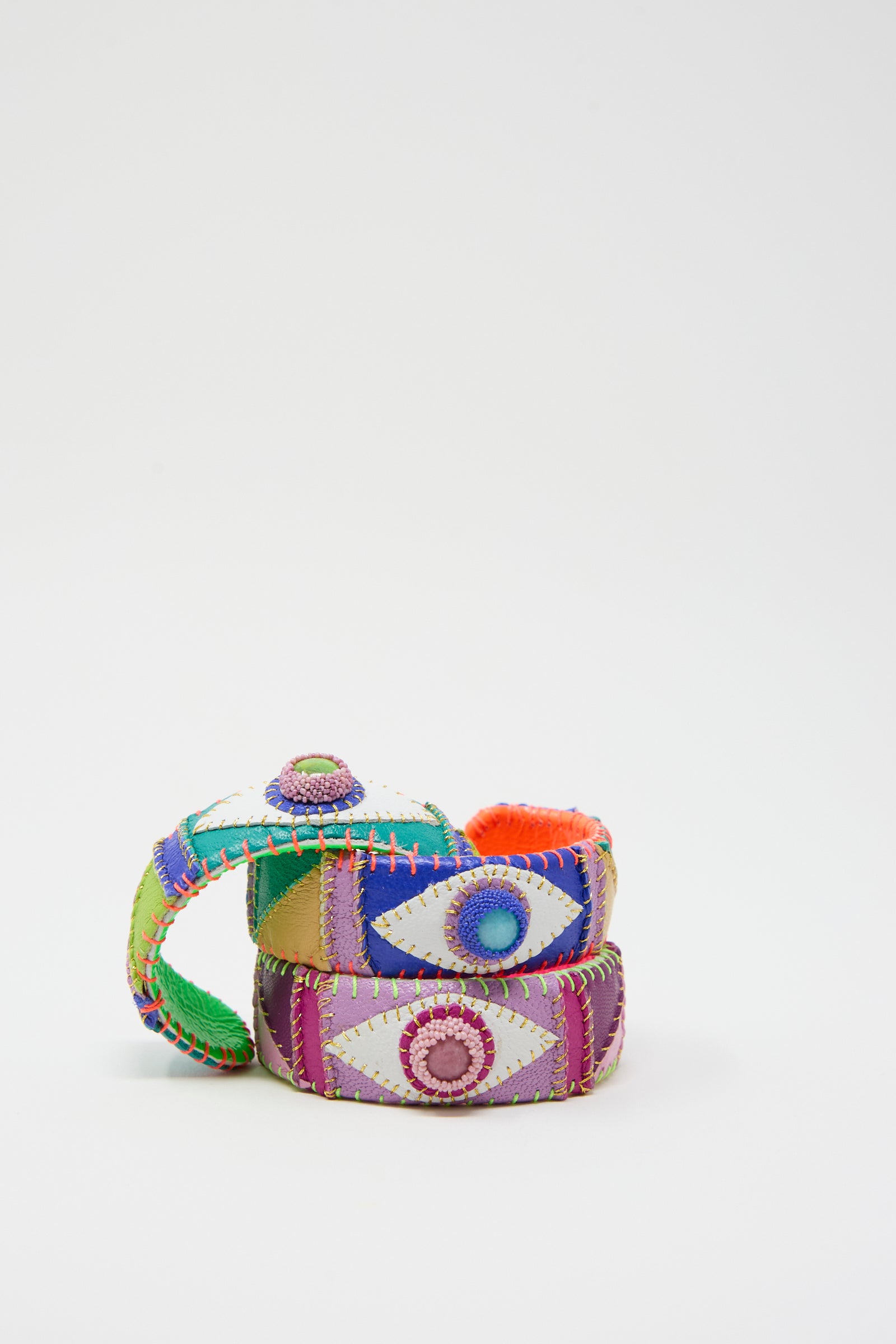 Three colorful, fabric-wrapped bracelets with stitched patterns and buttons, stacked in a pyramid formation on a plain white background, are complemented by the intricately detailed Eye Cuff in Amazonite from Robin Mollicone.