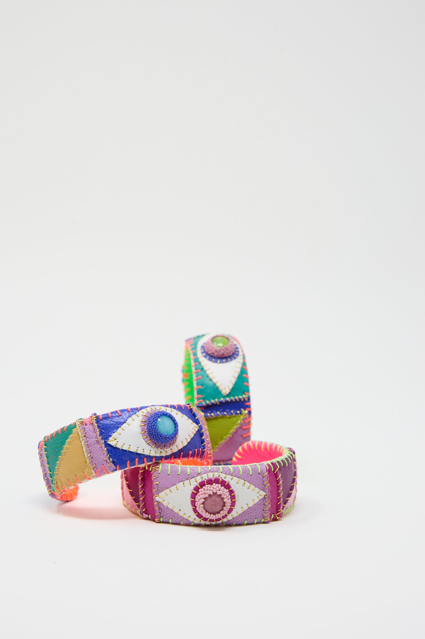 Three vibrant fabric bracelets called "Eye Cuff in Rhodonite" by Robin Mollicone, featuring eye designs of rhodonite stone, are arranged against a plain white background.