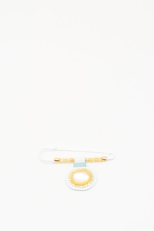 The Gem Drop Brooch in Mother of Pearl by Robin Mollicone features gold and white beads with a circular Mother of Pearl pendant, displayed on a plain white background.