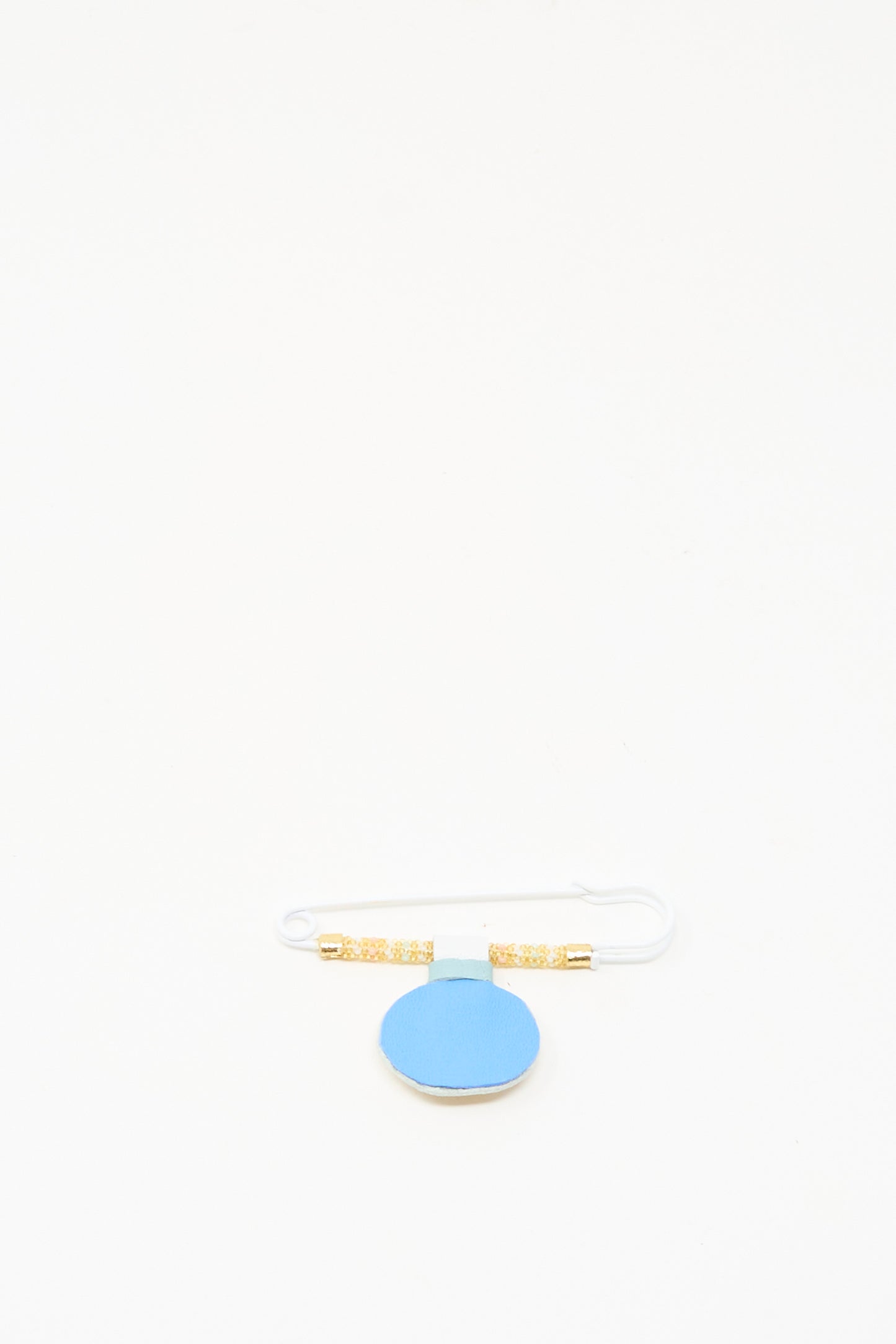 The Gem Drop Brooch in Mother of Pearl by Robin Mollicone features beaded safety pin details with gold accents on a round blue tag, displayed against a white background. Handmade in NYC, it brings elegance to any collection.