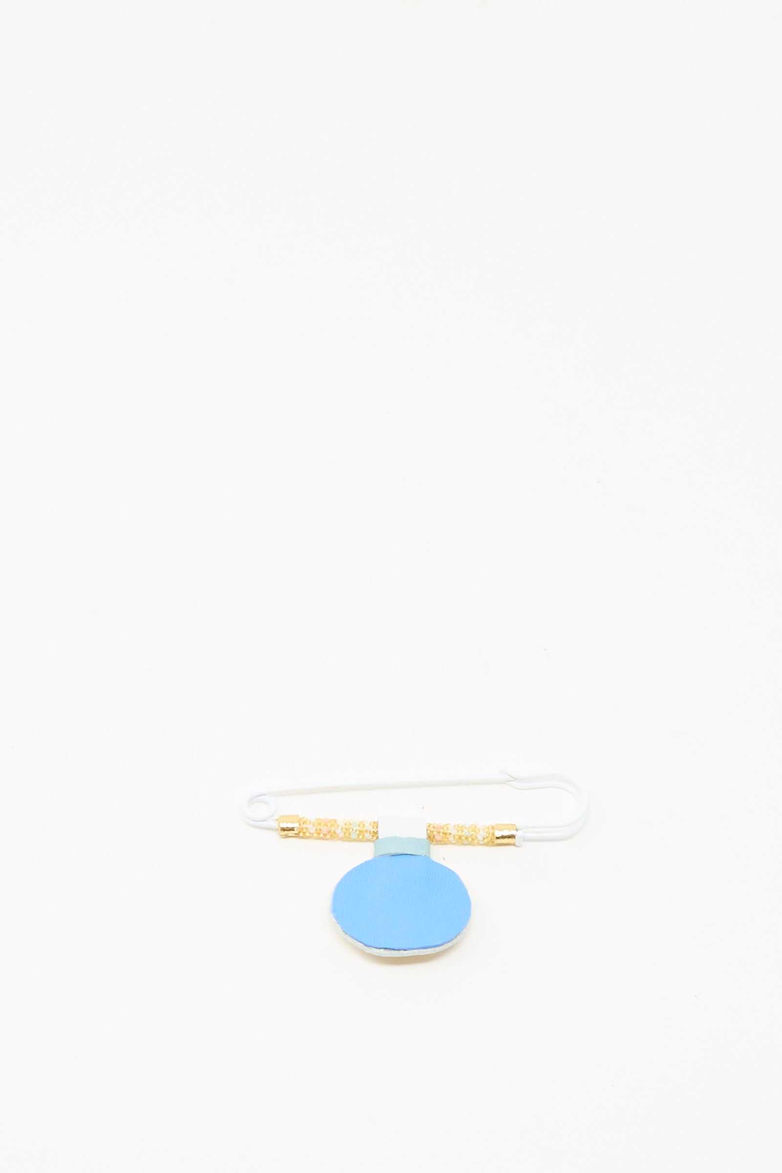 The Gem Drop Brooch in Mother of Pearl by Robin Mollicone features beaded safety pin details with gold accents on a round blue tag, displayed against a white background. Handmade in NYC, it brings elegance to any collection.
