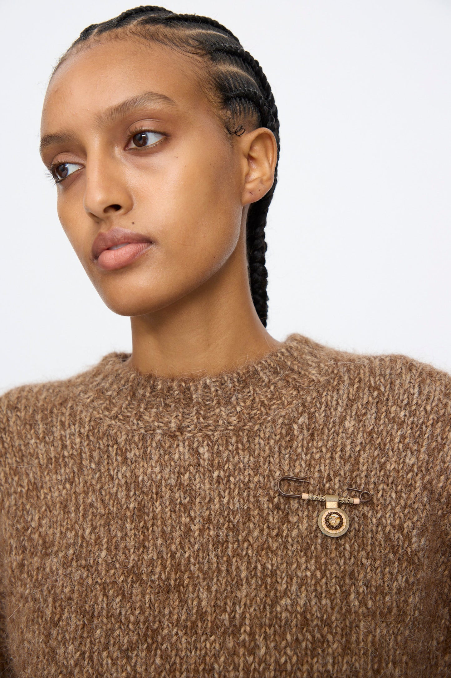 A person wears a chunky brown knit sweater adorned with the Robin Mollicone Gem Drop Safety Pin in Brown with Picture Jasper, handmade in NYC, on the left side; hair styled in cornrows.