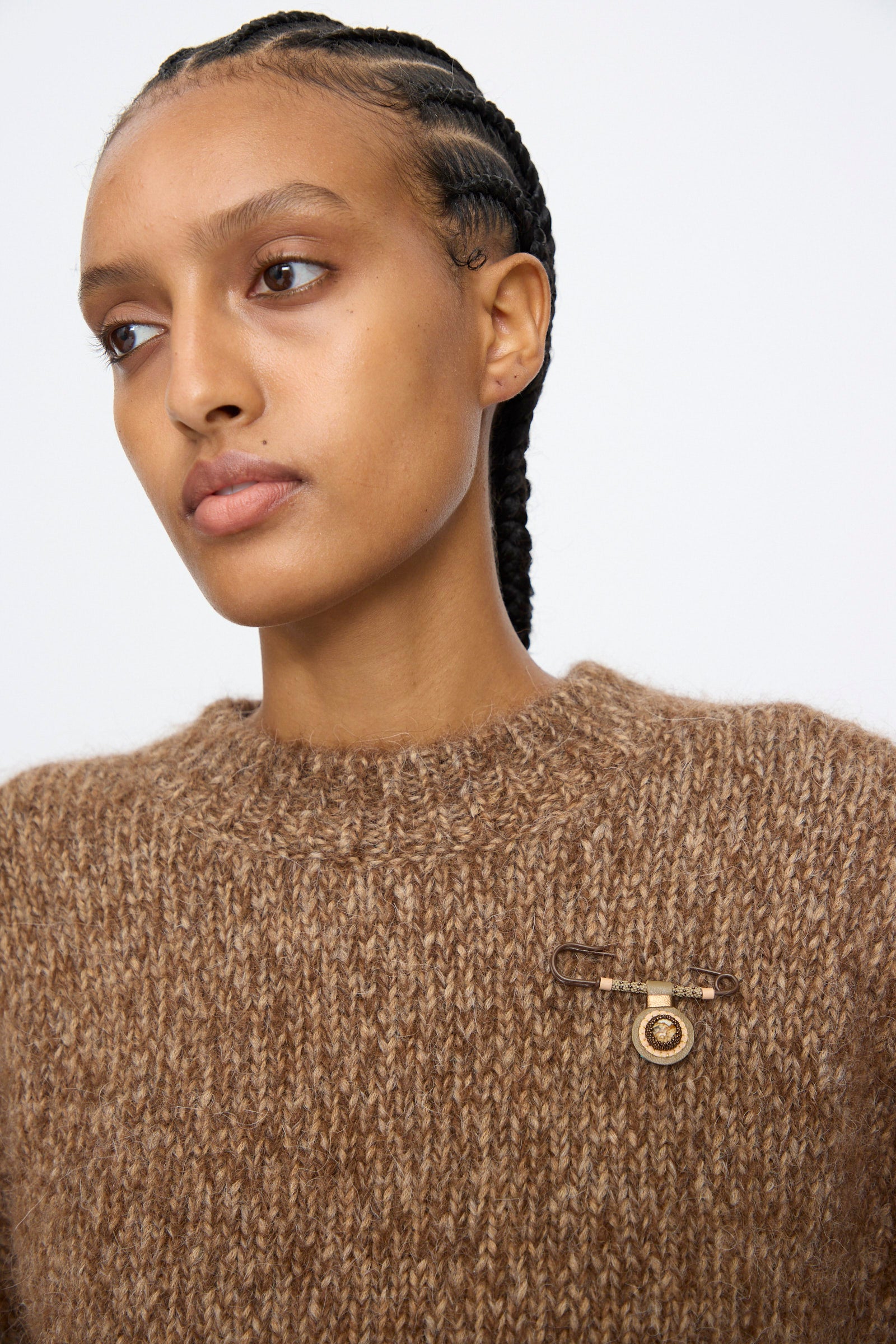 A person wears a chunky brown knit sweater adorned with the Robin Mollicone Gem Drop Safety Pin in Brown with Picture Jasper, handmade in NYC, on the left side; hair styled in cornrows.