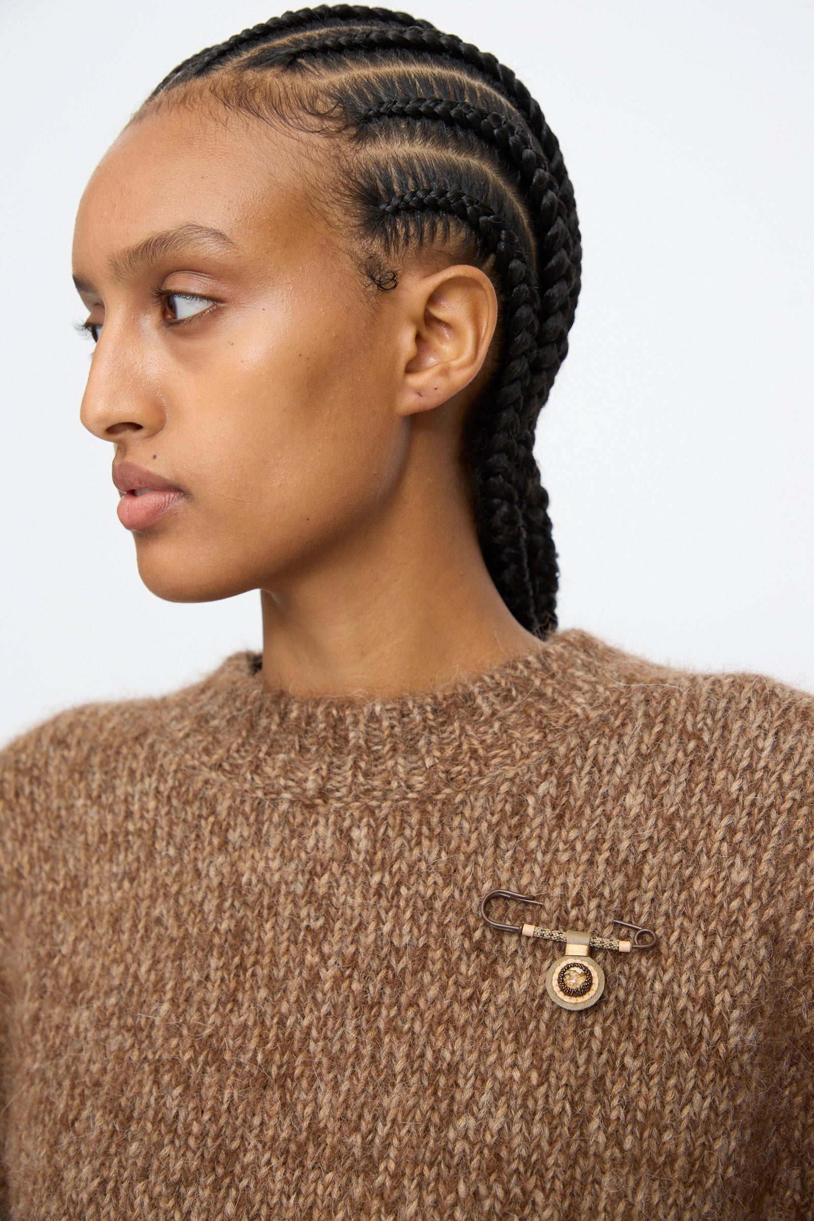 A person with braided hair wears a brown knit sweater adorned with the Robin Mollicone Gem Drop Safety Pin in Brown with Picture Jasper, handmade in NYC, on the left side.