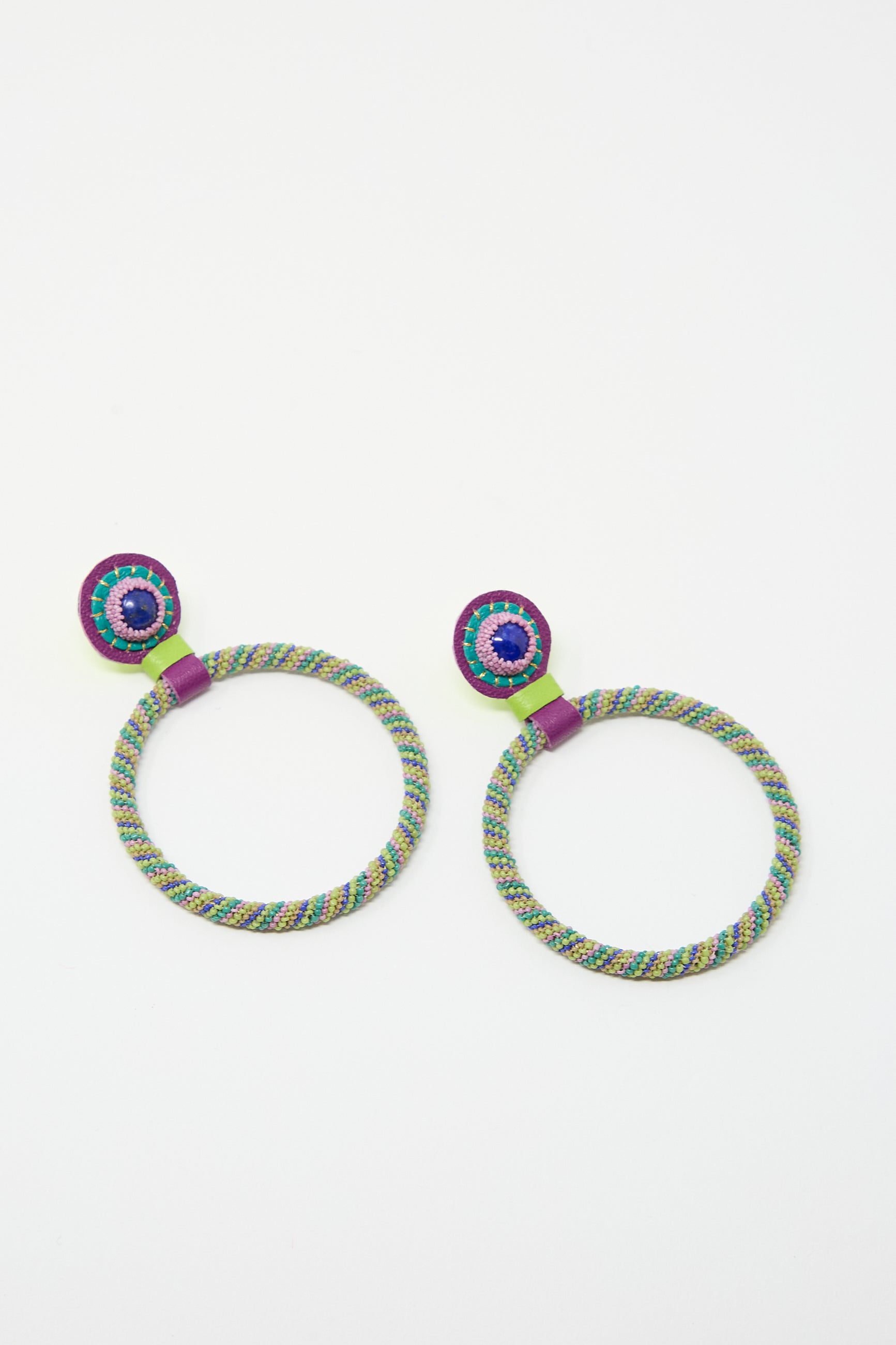 Two colorful, circular earrings with a woven texture and sterling silver posts, and a beaded embellishment at the top are placed side by side on a white background. These are the Large Beaded Hoop Earrings in Multi Spiral with Lapis Stone Tops by Robin Mollicone.