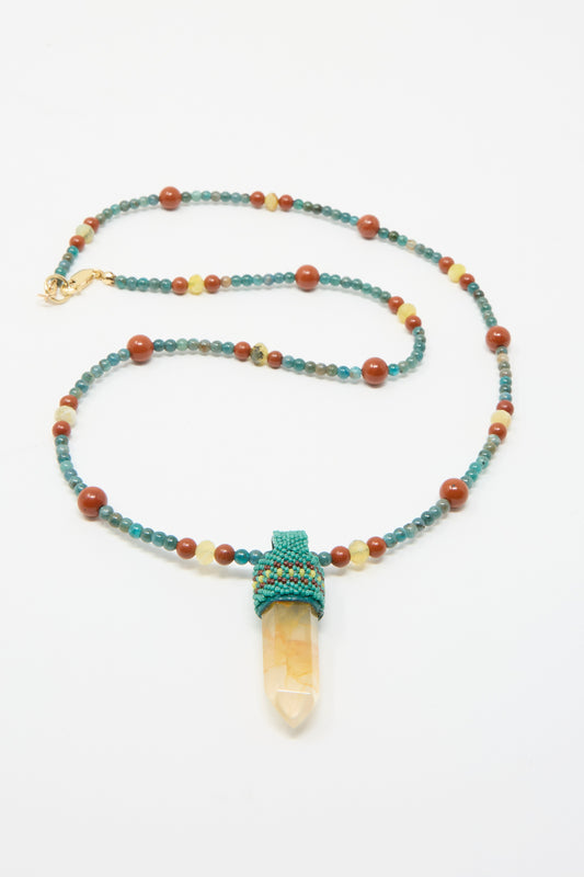 A Long Pendulum Necklace by Robin Mollicone features blue Apatite and brown Red Jasper beads, olive opal beads, and a clear quartz pendant with a woven holder, against a white background. 