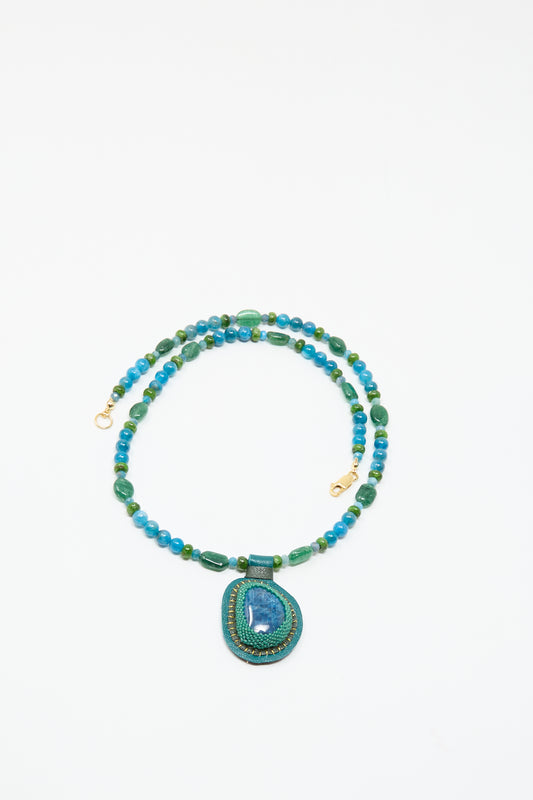The Love Bead Charm Necklace in Blue with Apatite Charm by Robin Mollicone features vintage blue and green glass beads, a round blue-green pendant, and gold clasp, shown on a white background.