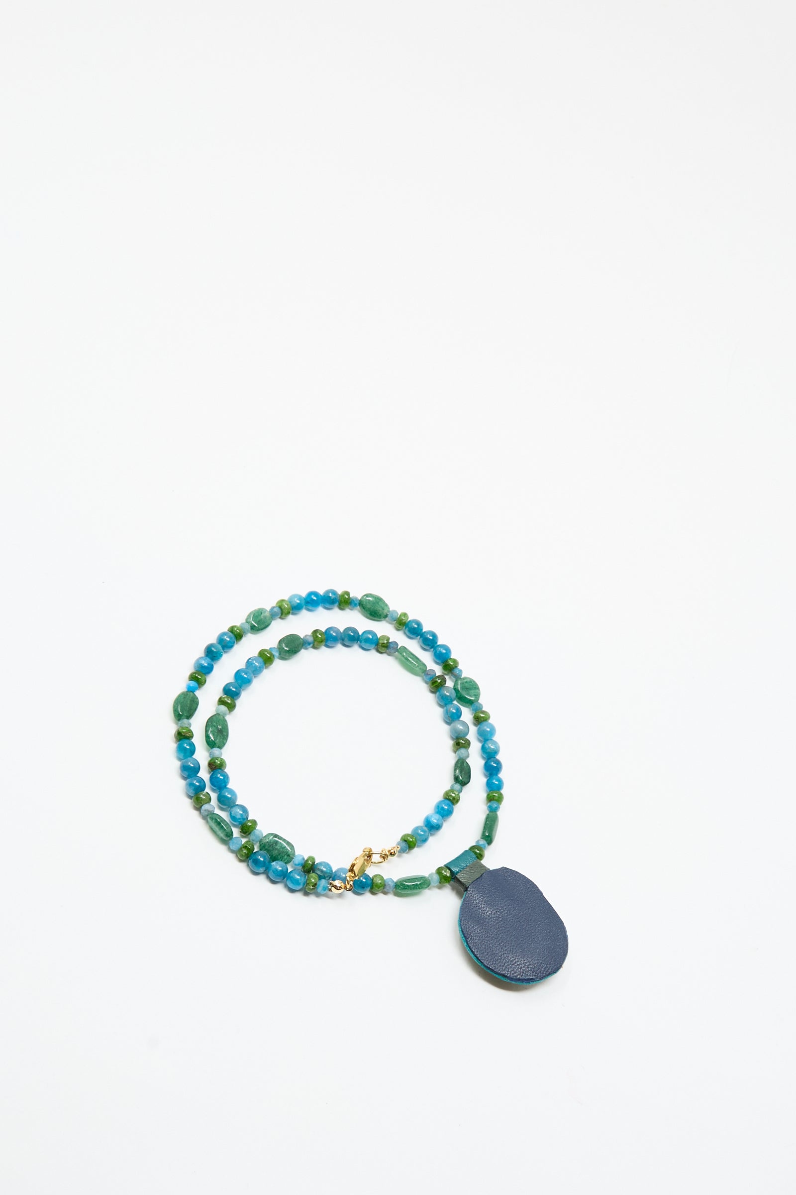 The Love Bead Charm Necklace in Blue with Apatite Charm by Robin Mollicone features vintage blue and green glass beads, a round navy pendant, and a gold clasp, displayed on a white surface.