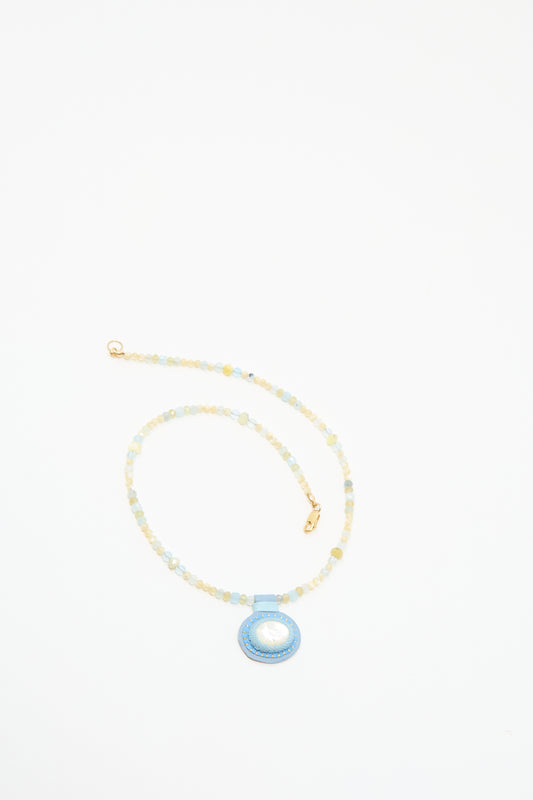 The Robin Mollicone Love Bead Charm Necklace Aquamarine with Mother Of Pearl Charm, featuring pale blue and cream vintage glass beads and a round blue pendant with a white Mother of Pearl center, is displayed on a white background.