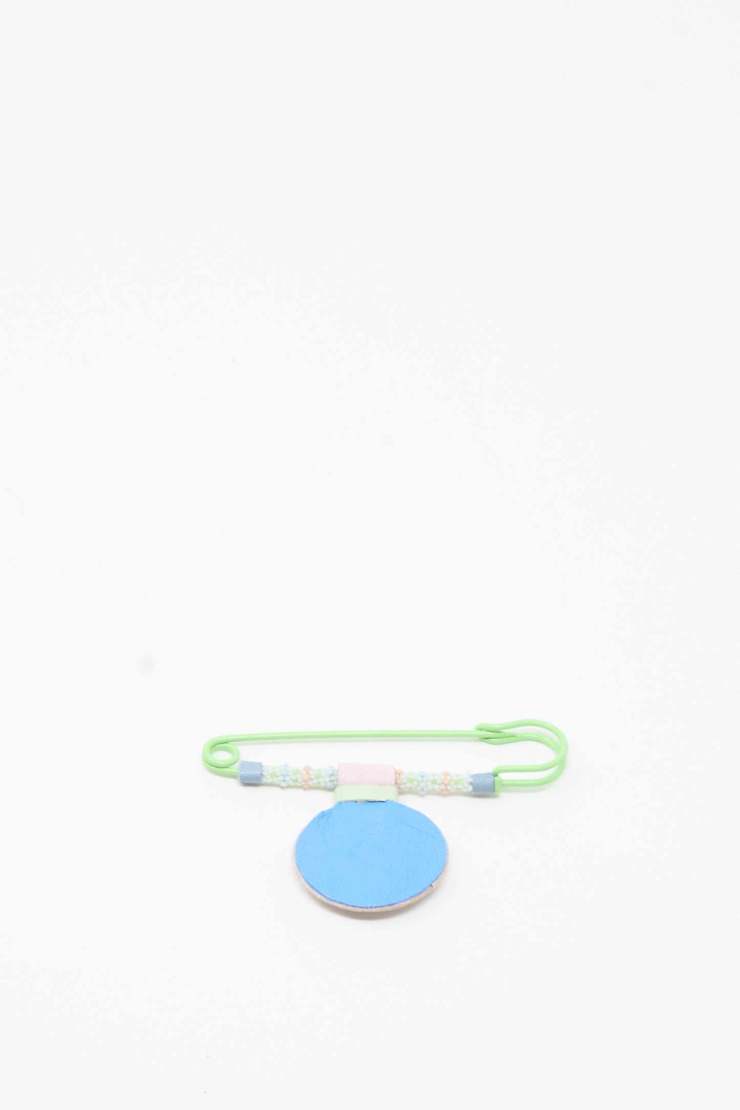 A handmade Robin Mollicone Mother of Pearl Gem Drop Brooch in pink, blue, and green is displayed on a round, flat blue surface against a white background.