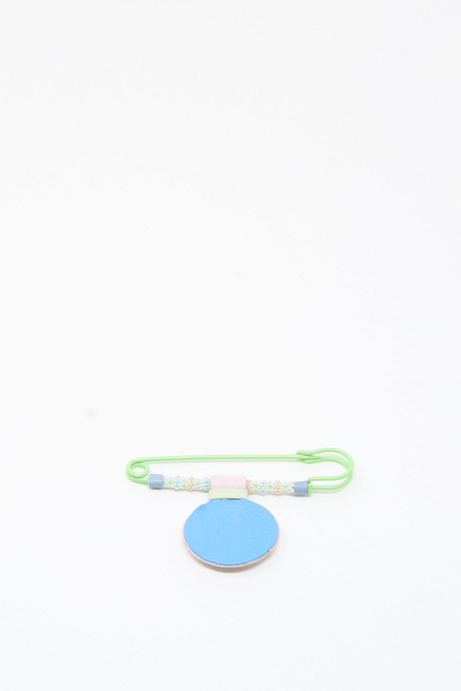 A handmade Robin Mollicone Mother of Pearl Gem Drop Brooch in pink, blue, and green is displayed on a round, flat blue surface against a white background.
