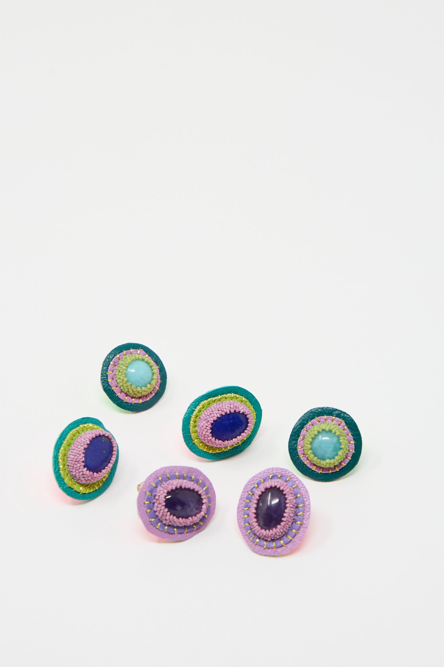 Six multicolored, oval-shaped "Oval Earrings in Amethyst" by Robin Mollicone are arranged on a white background. Each earring features a colorful bead in the center, surrounded by intricate threadwork reminiscent of the detail found in handmade brooches.