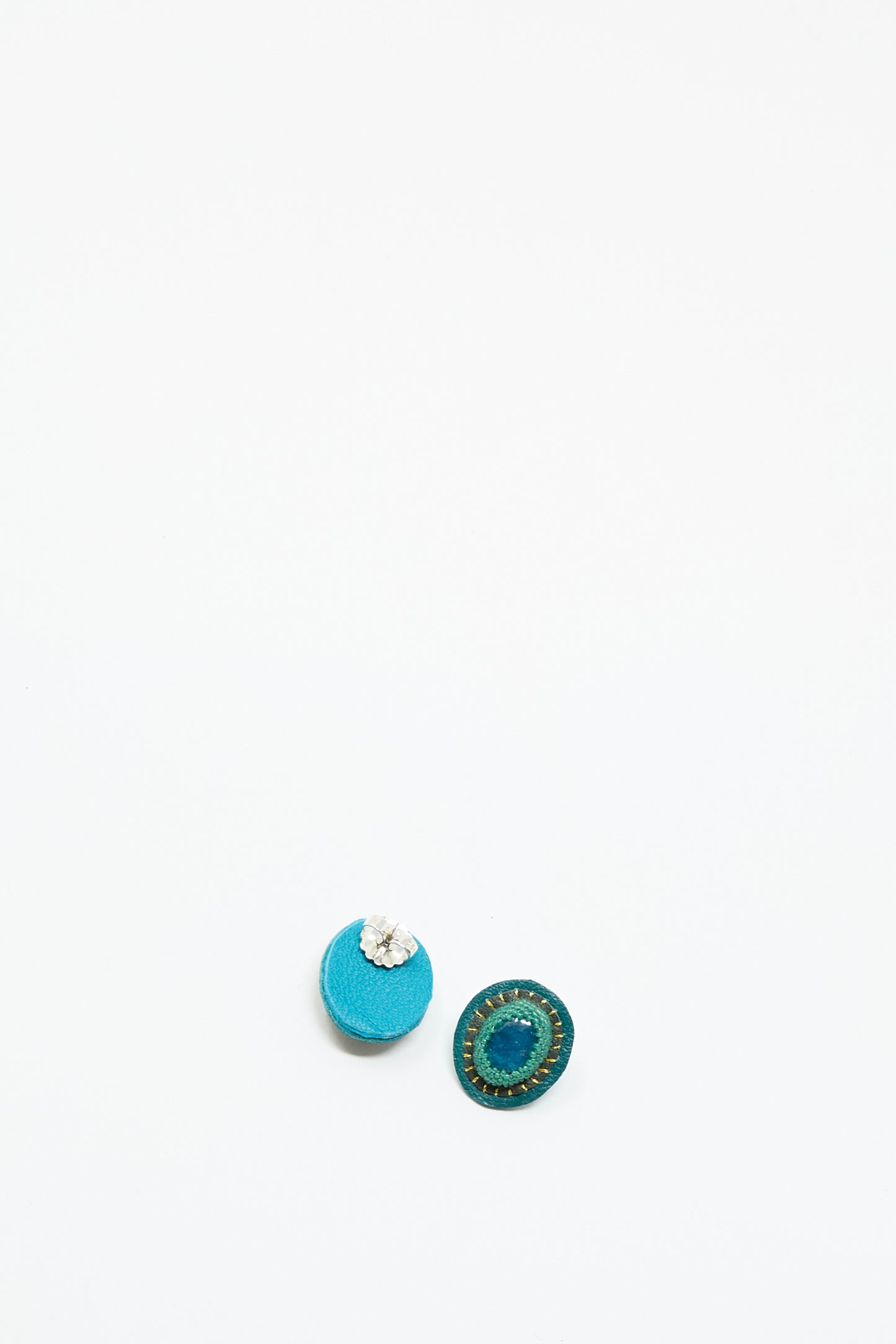 The Oval Earrings in Apatite by Robin Mollicone are displayed on a plain white background, featuring one blue with silver detail and one dark green with a stitched pattern, each handmade for a unique look.