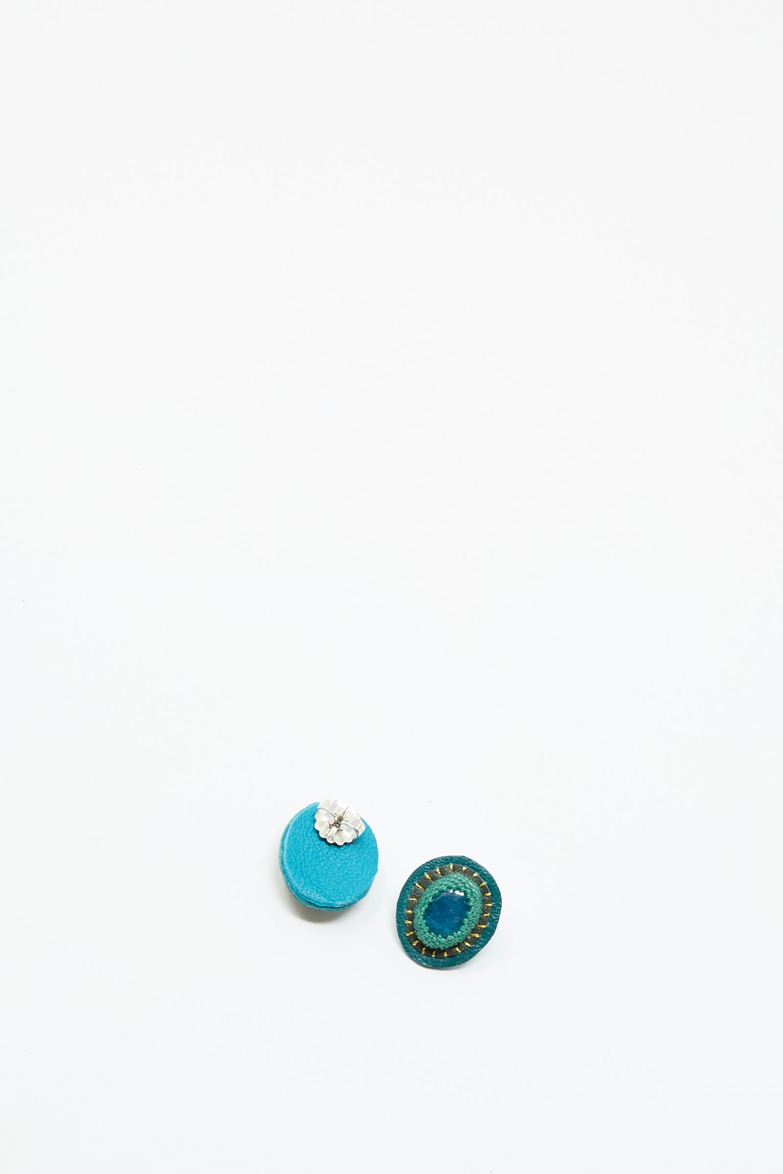 The Oval Earrings in Apatite by Robin Mollicone are displayed on a plain white background, featuring one blue with silver detail and one dark green with a stitched pattern, each handmade for a unique look.