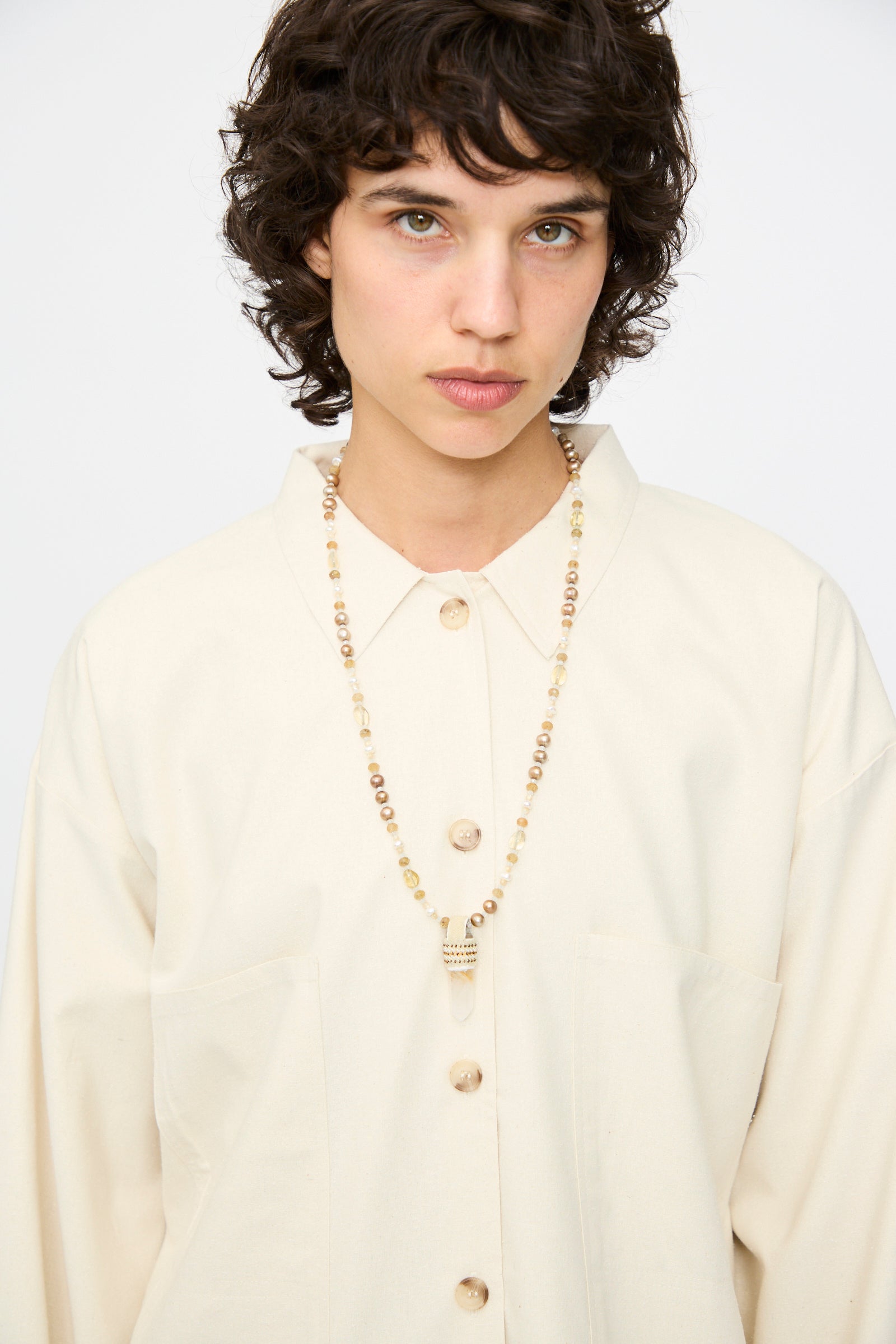 Person with short curly hair wears a cream button-up shirt and the Robin Mollicone Peace of Mind Necklace, featuring mother of pearl, vintage glass beads, and a quartz crystal pendant, standing against a plain light background.