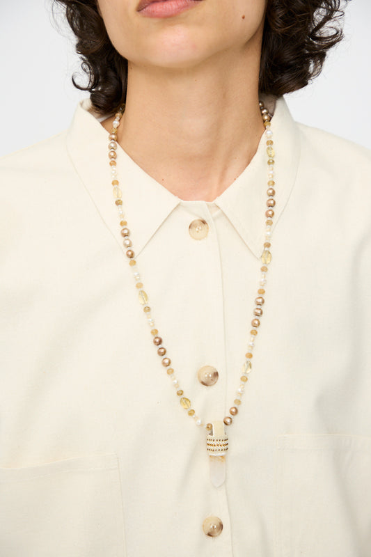 A person wearing a cream button-up shirt and the Robin Mollicone Peace of Mind Necklace in Mother of Pearl with Quartz Crystal Pendant, photographed from the shoulders up.