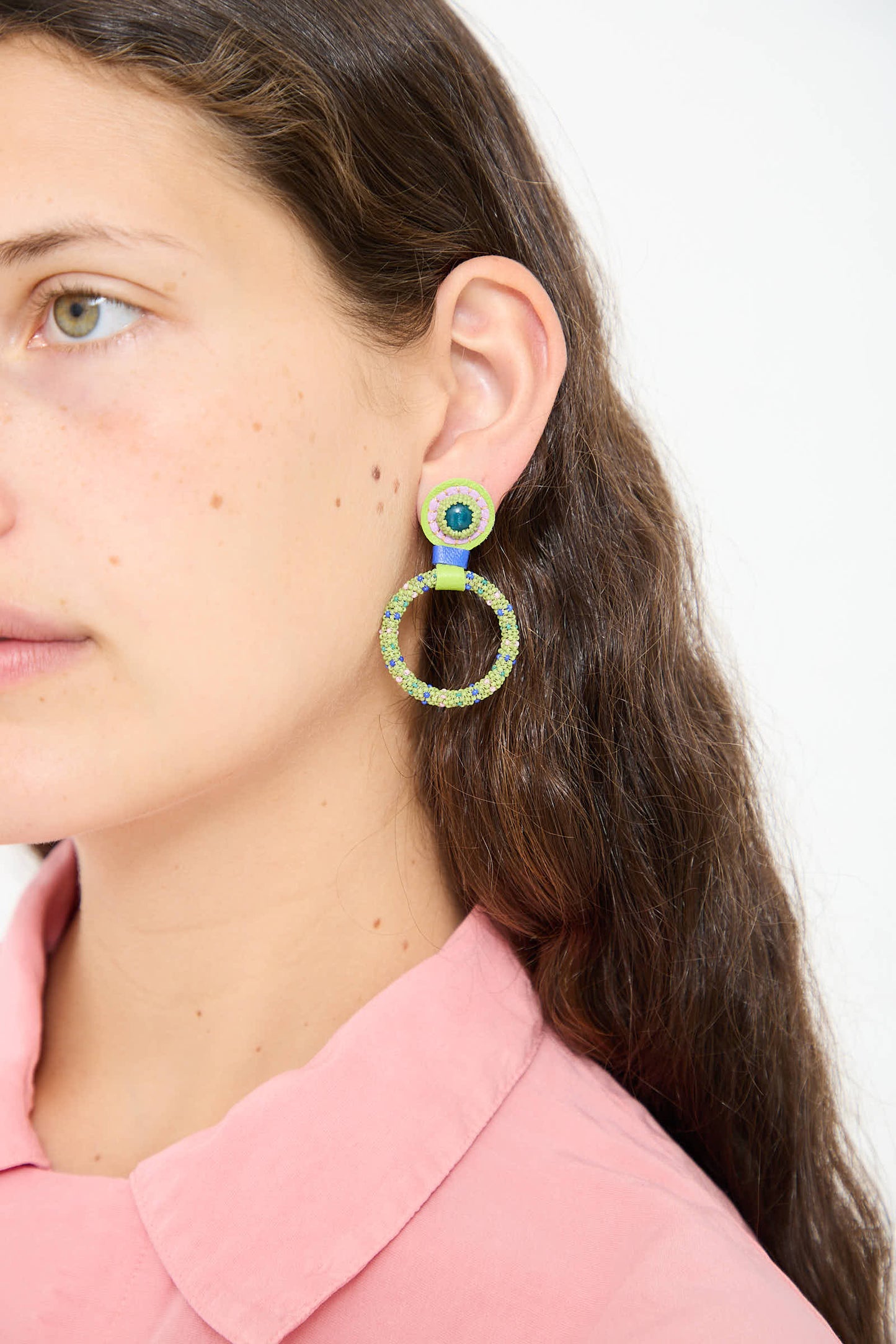 Close-up of a person with long brown hair wearing Robin Mollicone Small Beaded Hoop Earrings in Olive Dots with Apatite Stone Tops, dressed in a pink collared shirt against a plain white background.