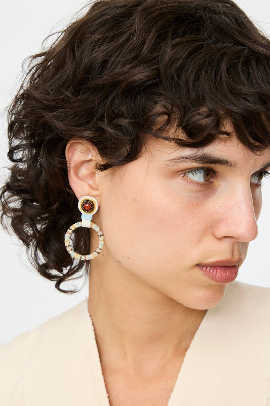 A woman with short, curly brown hair wears Robin Mollicone's Small Beaded Hoop Earrings in Red Jasper and a beige top, her neutral expression focused to the left.