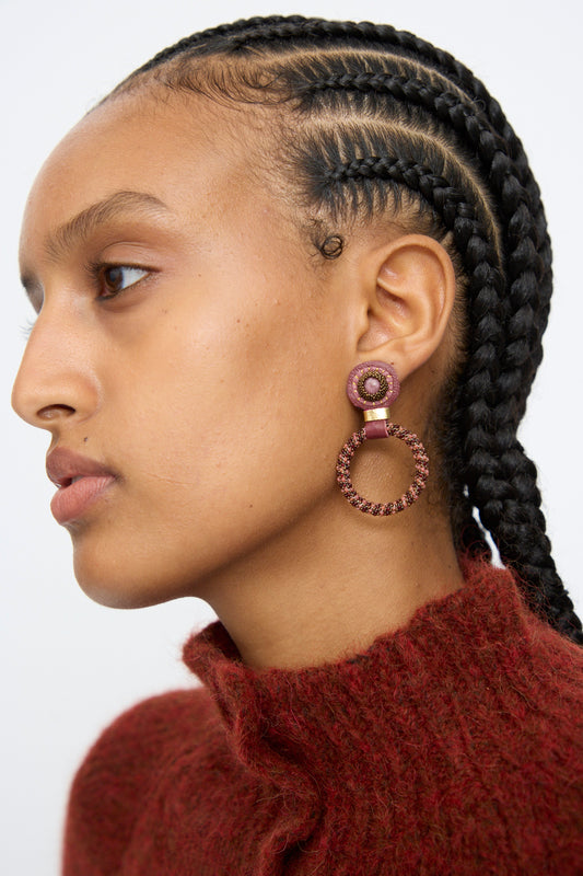 A person with braided hair wears a maroon textured sweater and Robin Mollicone's Small Beaded Hoops in Pink with Rhodonite, looking to the left against a plain background.
