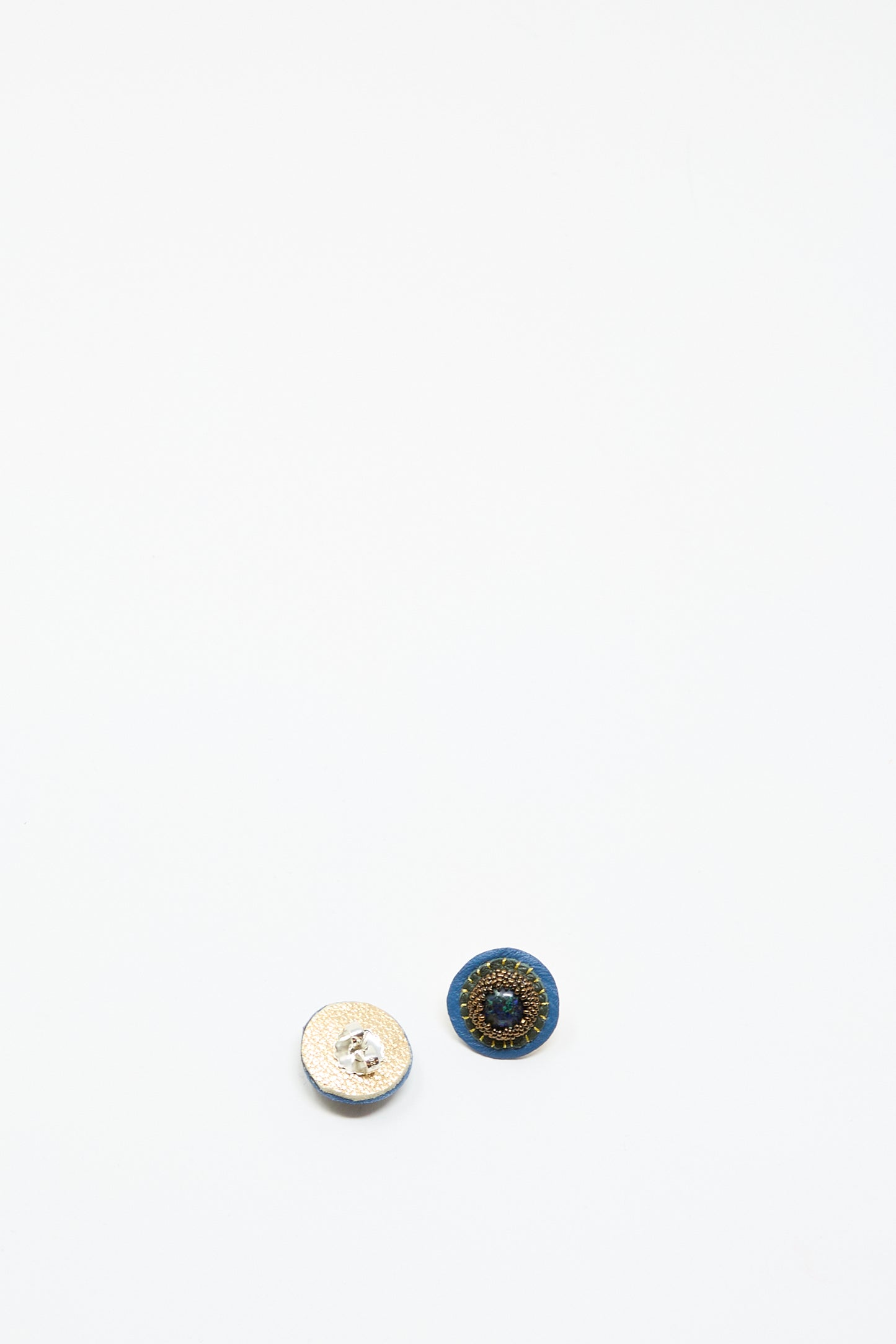 Two small circular buttons, one white textured and one dark with a blue border and gold center, are displayed on white—evoking the look of Robin Mollicone's Stud Earrings in Blue with Azurite.