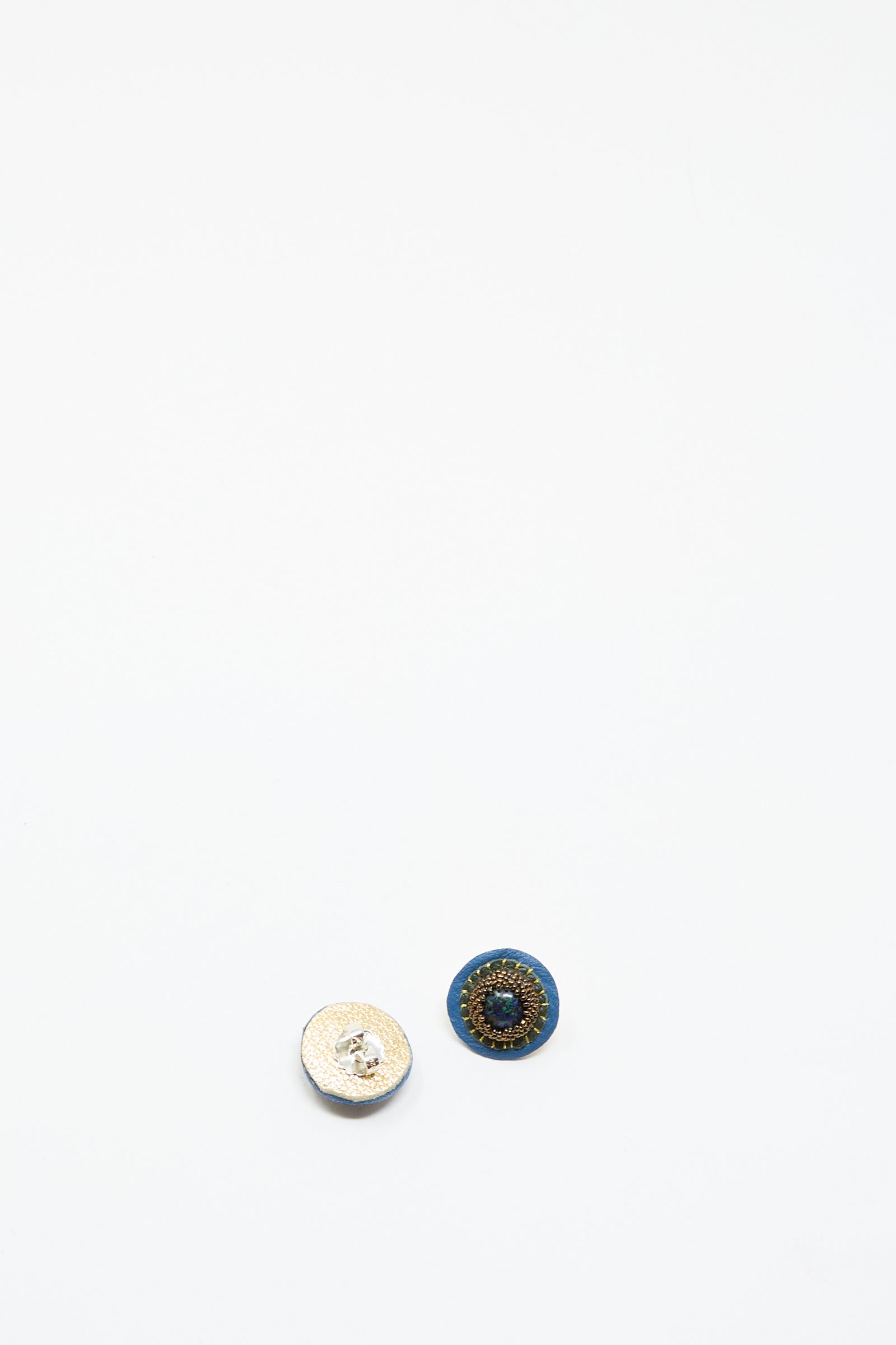 Two small circular buttons, one white textured and one dark with a blue border and gold center, are displayed on white—evoking the look of Robin Mollicone's Stud Earrings in Blue with Azurite.