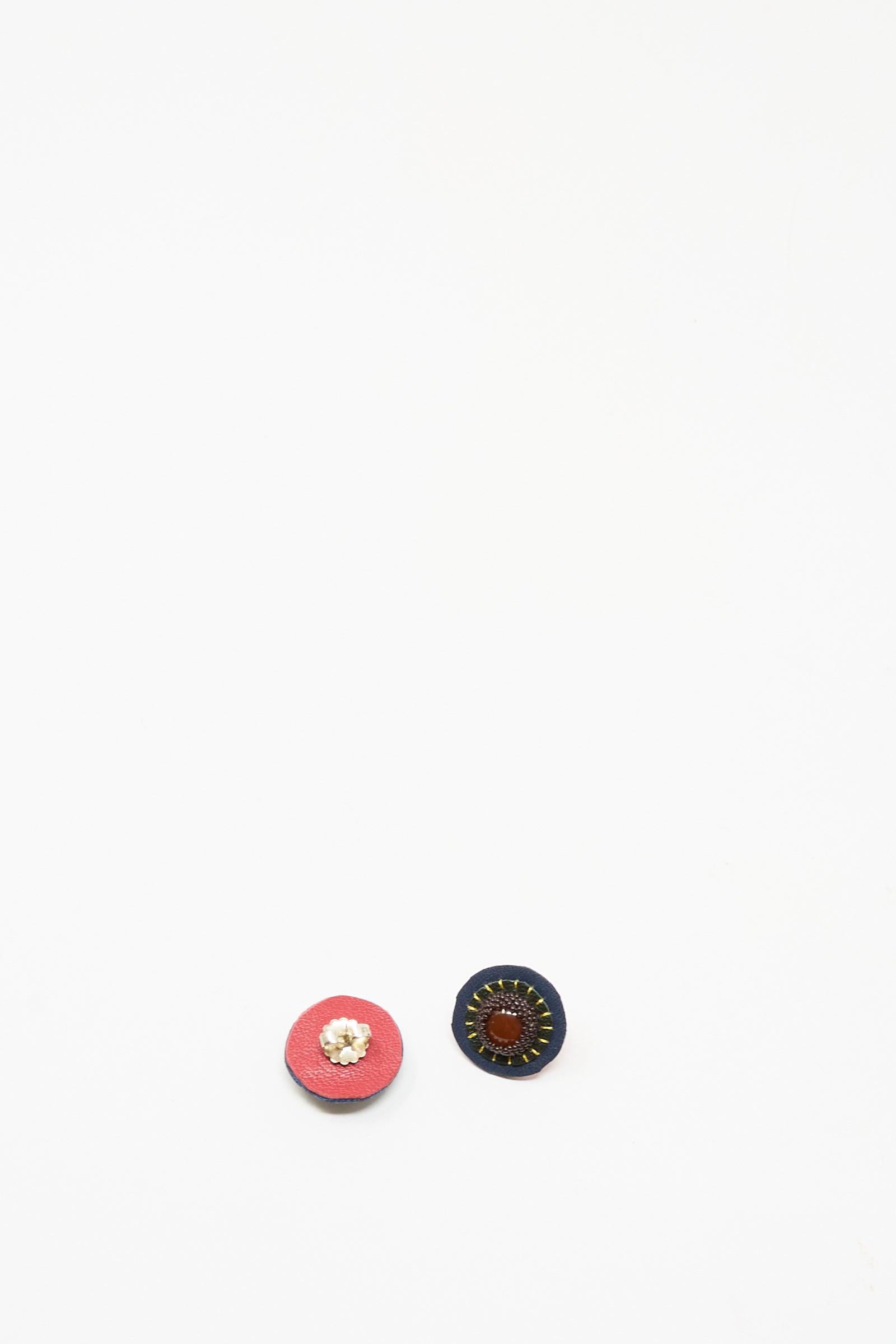 Two round stud earrings—one pink with a floral motif and one dark blue with stitched detail from Robin Mollicone’s Carnelian collection—are displayed on a plain white background, exuding handmade charm.