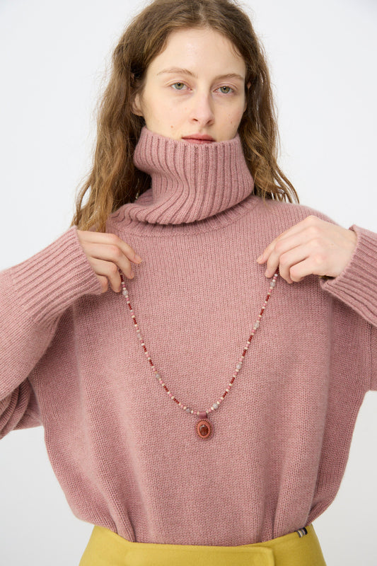 A person in a chunky pink turtleneck and yellow pants holds the Robin Mollicone Love Bead Necklace in Pink with Carnelian Charm, featuring vintage glass beads and a circular pendant, handmade in NYC.