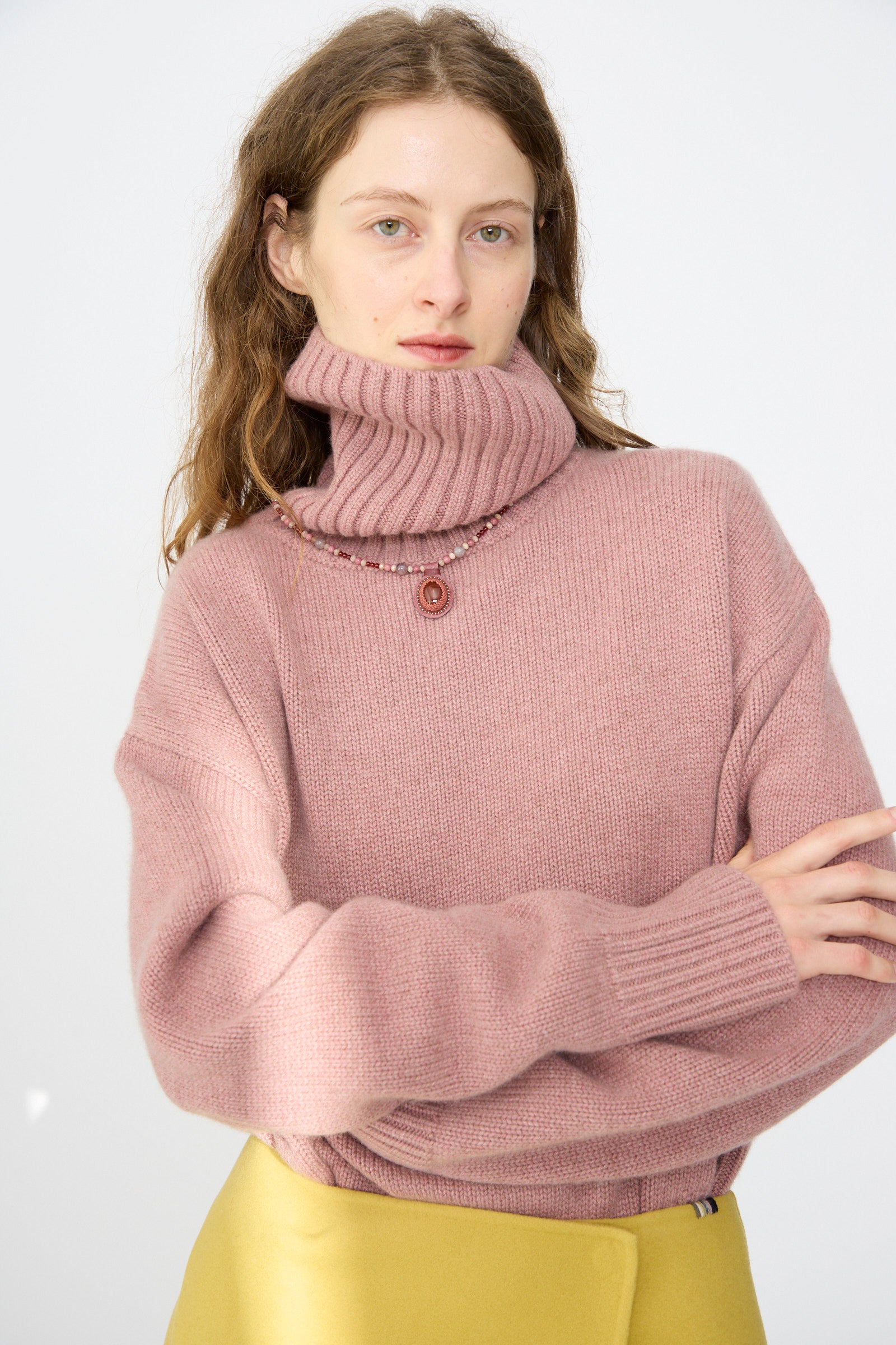 A woman with wavy brown hair wears a pink turtleneck sweater, a yellow skirt, and the Robin Mollicone Love Bead Necklace in Pink with Carnelian Charm, standing against a plain white background.