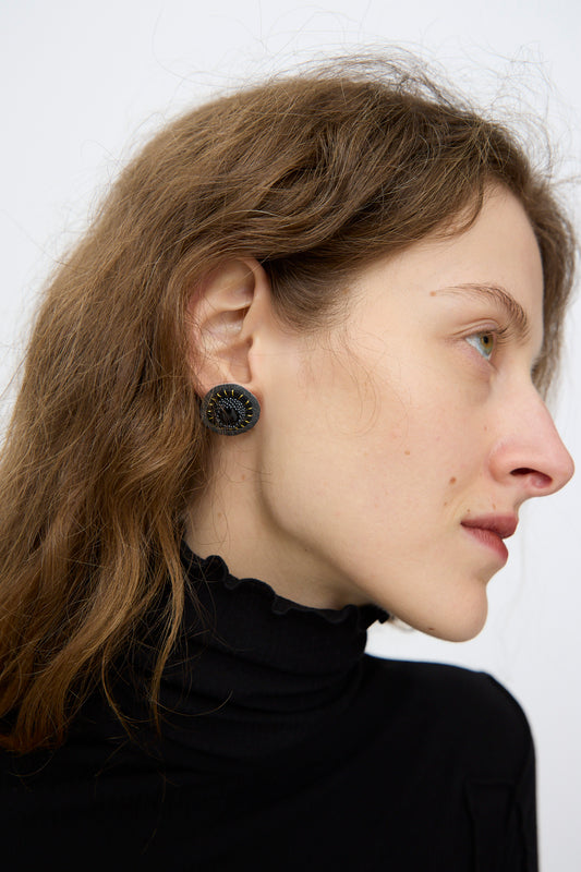 A person with wavy brown hair wears a black high-neck top and Robin Mollicone's Stud Earrings in Onyx, shown in profile against a plain background.