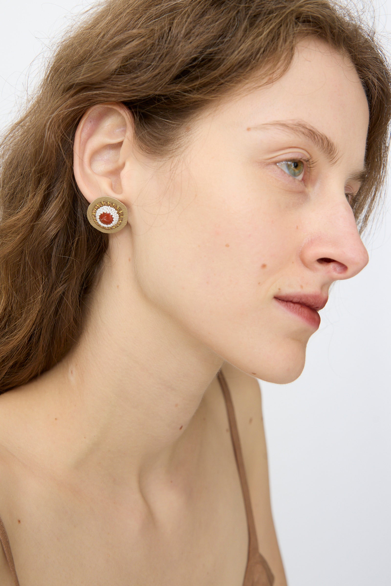 A light-skinned woman with brown hair is shown in profile wearing Robin Mollicone's Stud Earrings in Red Jasper, featuring a red center and beige outer ring. She wears a thin-strapped top against a plain background.