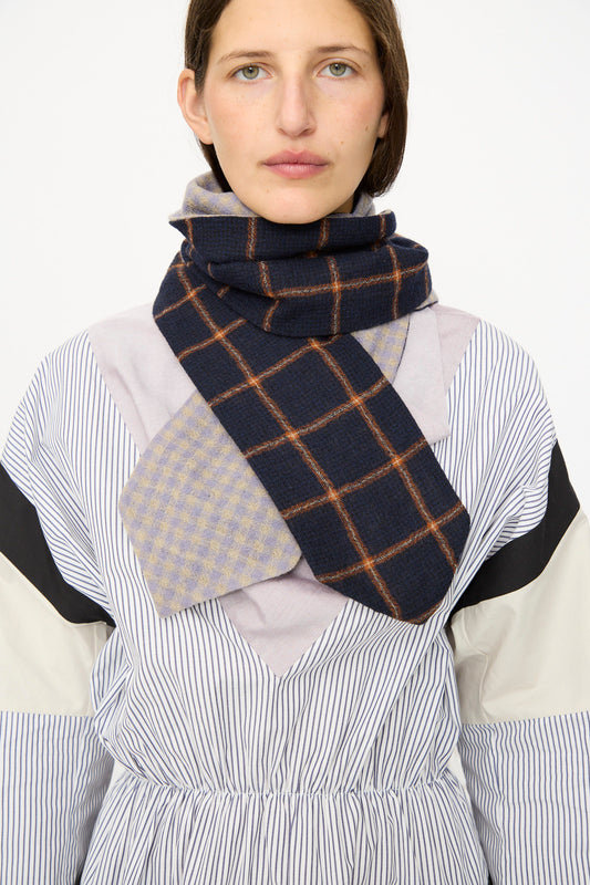 A person wears a striped dress paired with the SC103 Check Wool Latch Scarf in Glossary, featuring blue plaid and light checkered designs, wrapped around their neck.