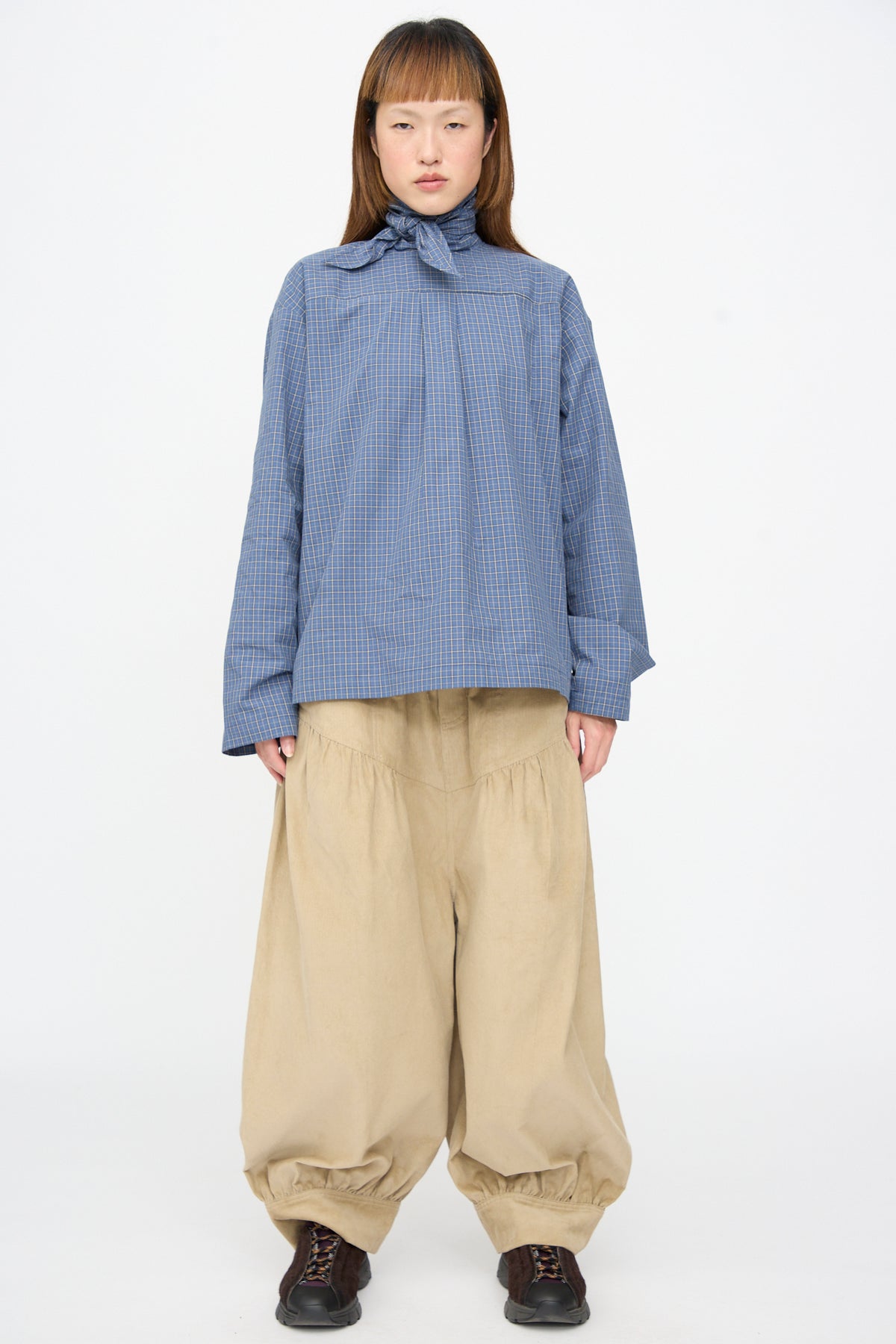 A person wears a blue checkered shirt with a neck tie and SC103 Cotton Corduroy Crescent Pants in Wheat with a balloon leg.