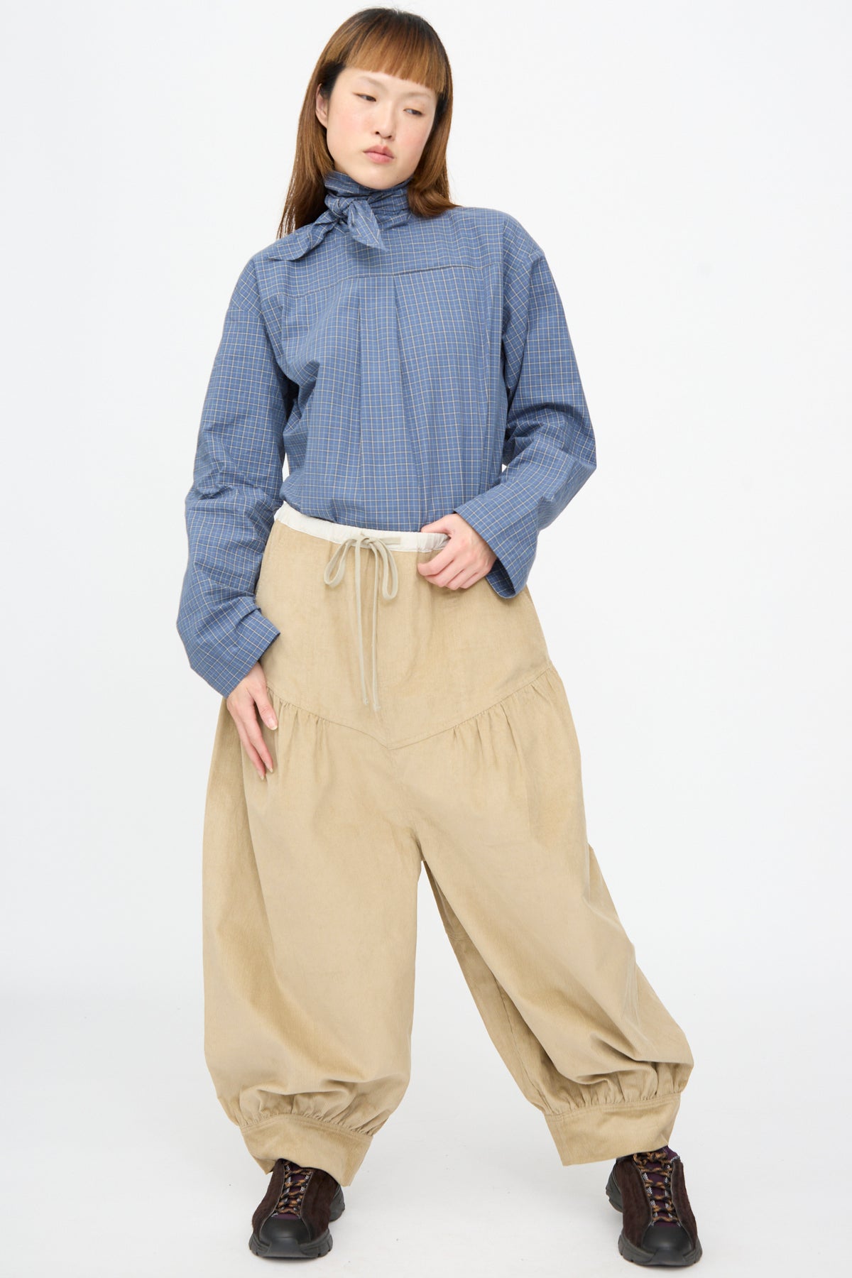 A person wearing a blue plaid long-sleeved shirt with a neck tie, beige oversized Cotton Corduroy Crescent Pants in Wheat, and dark sneakers.