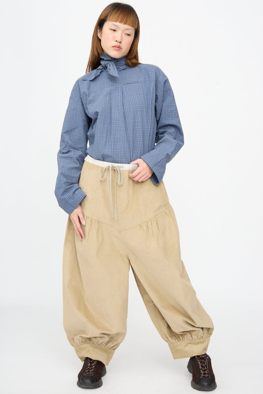 A person wearing a blue plaid long-sleeved shirt with a neck tie, beige oversized Cotton Corduroy Crescent Pants in Wheat, and dark sneakers.