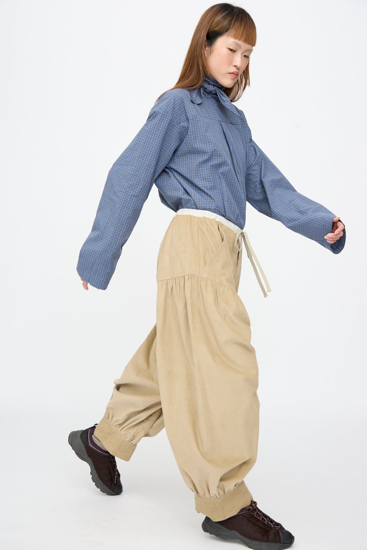 A person wears a blue plaid long-sleeved shirt and SC103 Cotton Corduroy Crescent Pants in Wheat, featuring an oversized balloon shape with an elastic waist and cuffs, paired with dark sneakers.