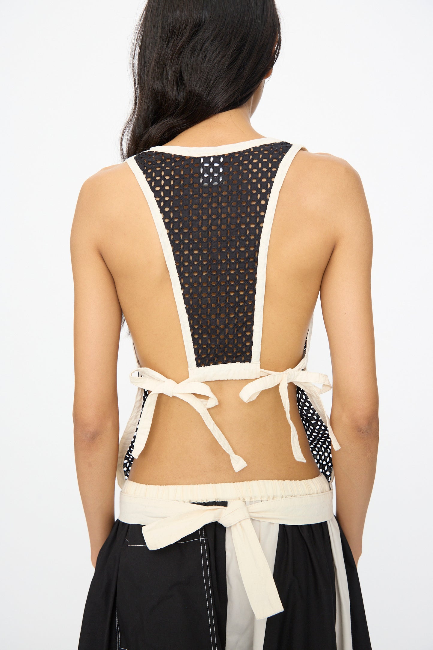 A woman faces away in the SC103 Cotton Eyelet Gaze Tank in Eclipse—a sleeveless black mesh top with cream trim and back ties—paired with a tie-waist skirt; thoughtfully crafted in NYC.