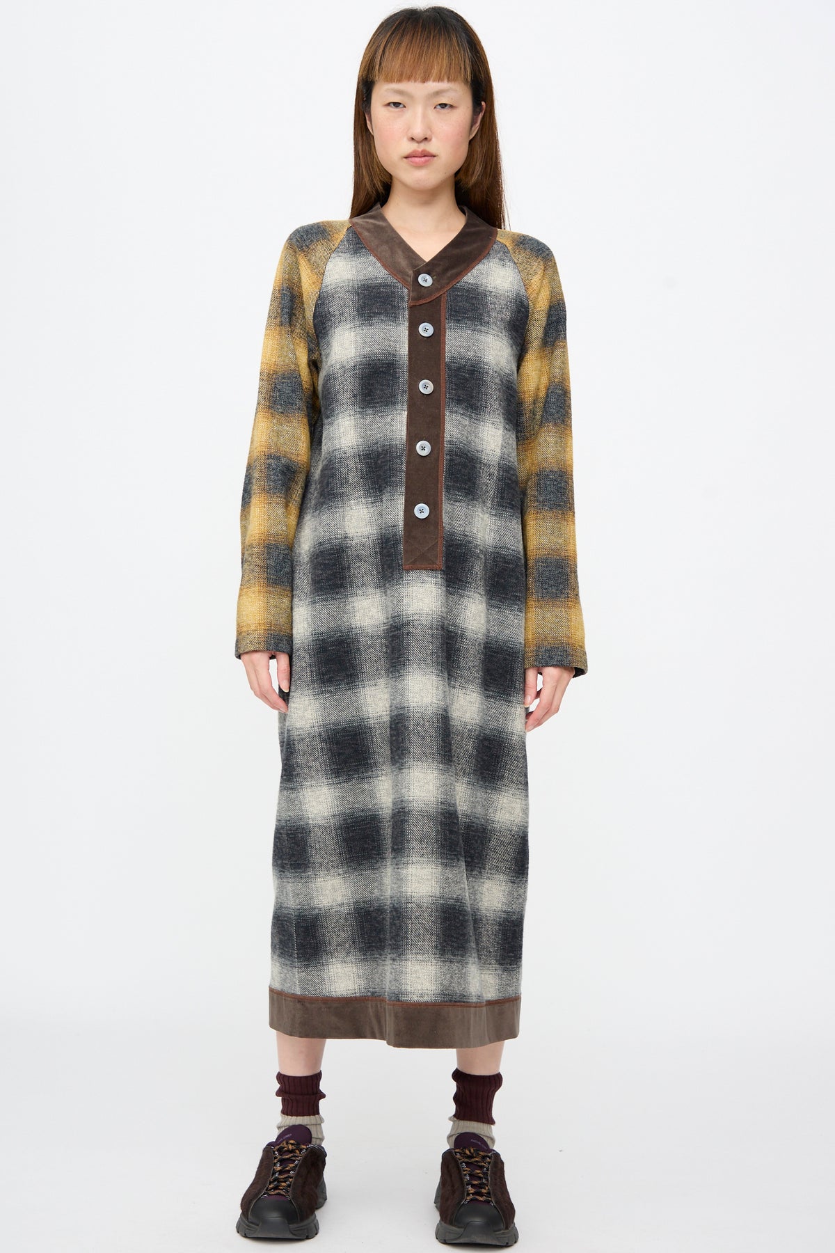 A person wearing a Cotton Flannel Frame Dress in Scholar, a long-sleeved, plaid midi dress with brown trim at the collar and hem, styled with dark sneakers and ribbed socks.