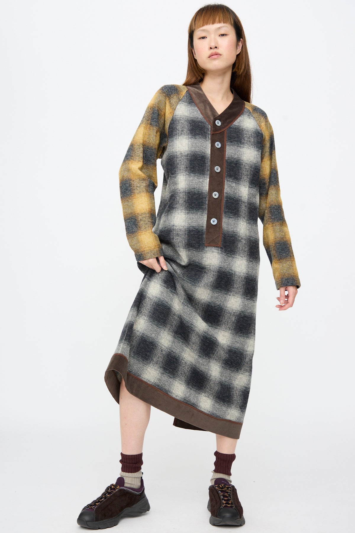 A model wears a SC103 Cotton Flannel Frame Dress in Scholar, a long-sleeved midi dress with a dark brown V-neck placket and grey and yellow plaid, paired with dark sneakers and maroon socks.