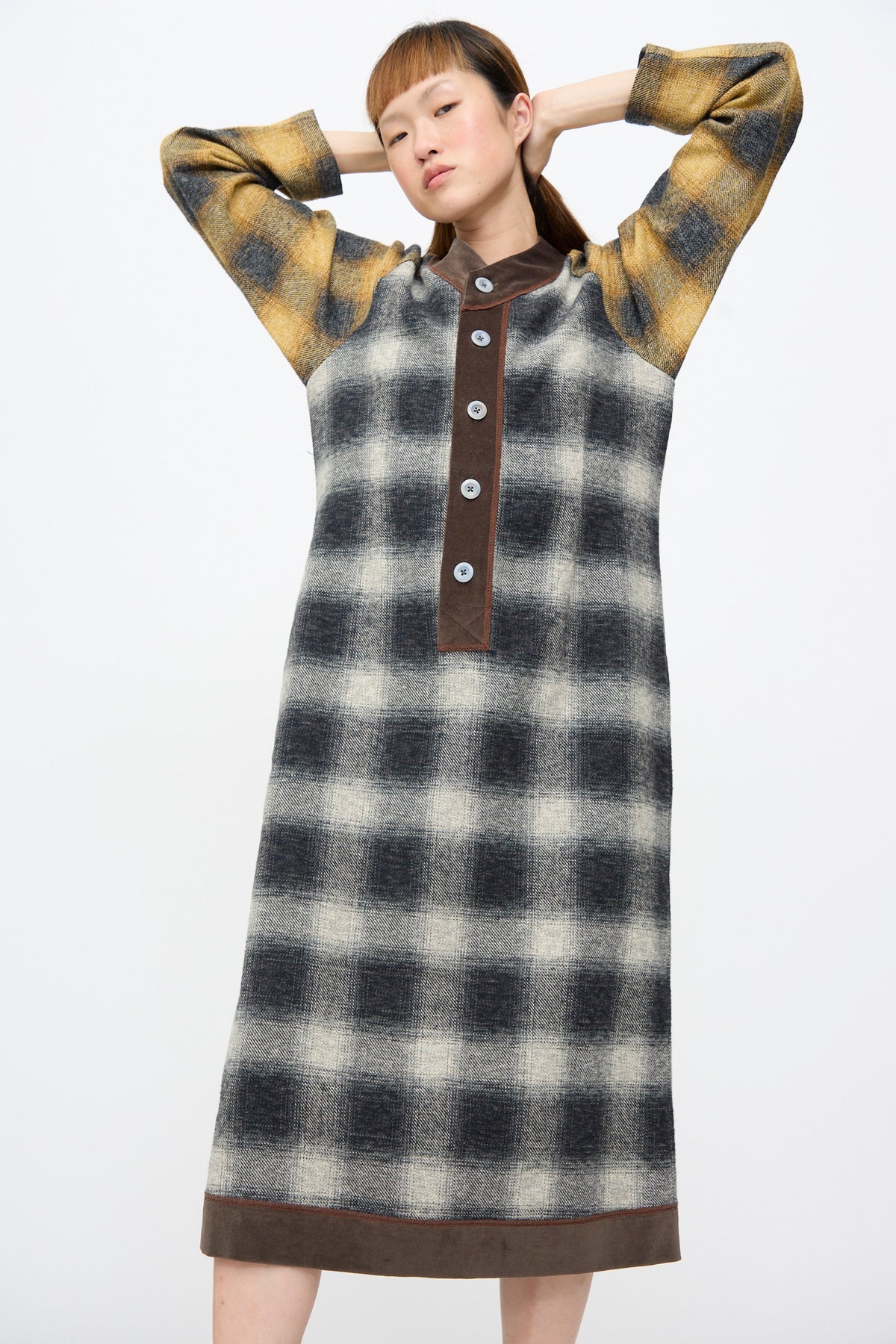 A person poses with hands behind their head wearing a Cotton Flannel Frame Dress in Scholar, featuring a black and white plaid pattern, brown trim, and a button-up placket.