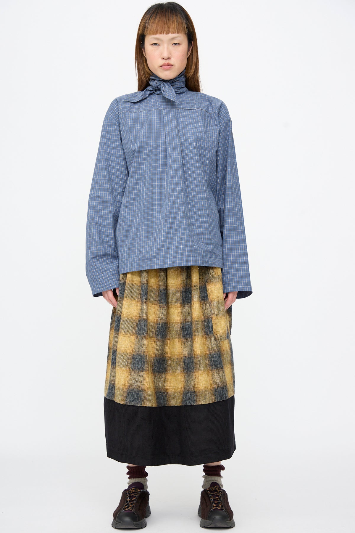 A person wearing a blue checkered blouse with a tied collar, a Cotton Flannel Rubric Skirt in Hearth, and dark sneakers.
