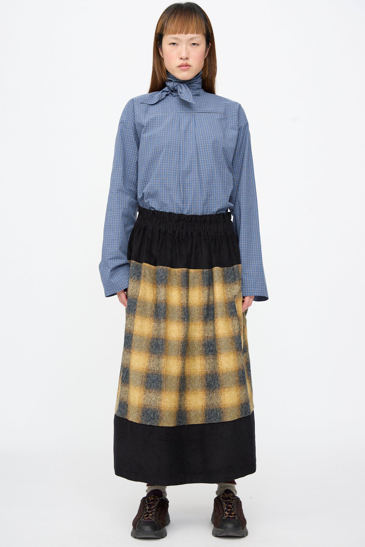 A person wearing a blue checkered shirt with a neck tie, and the Cotton Flannel Rubric Skirt in Hearth.